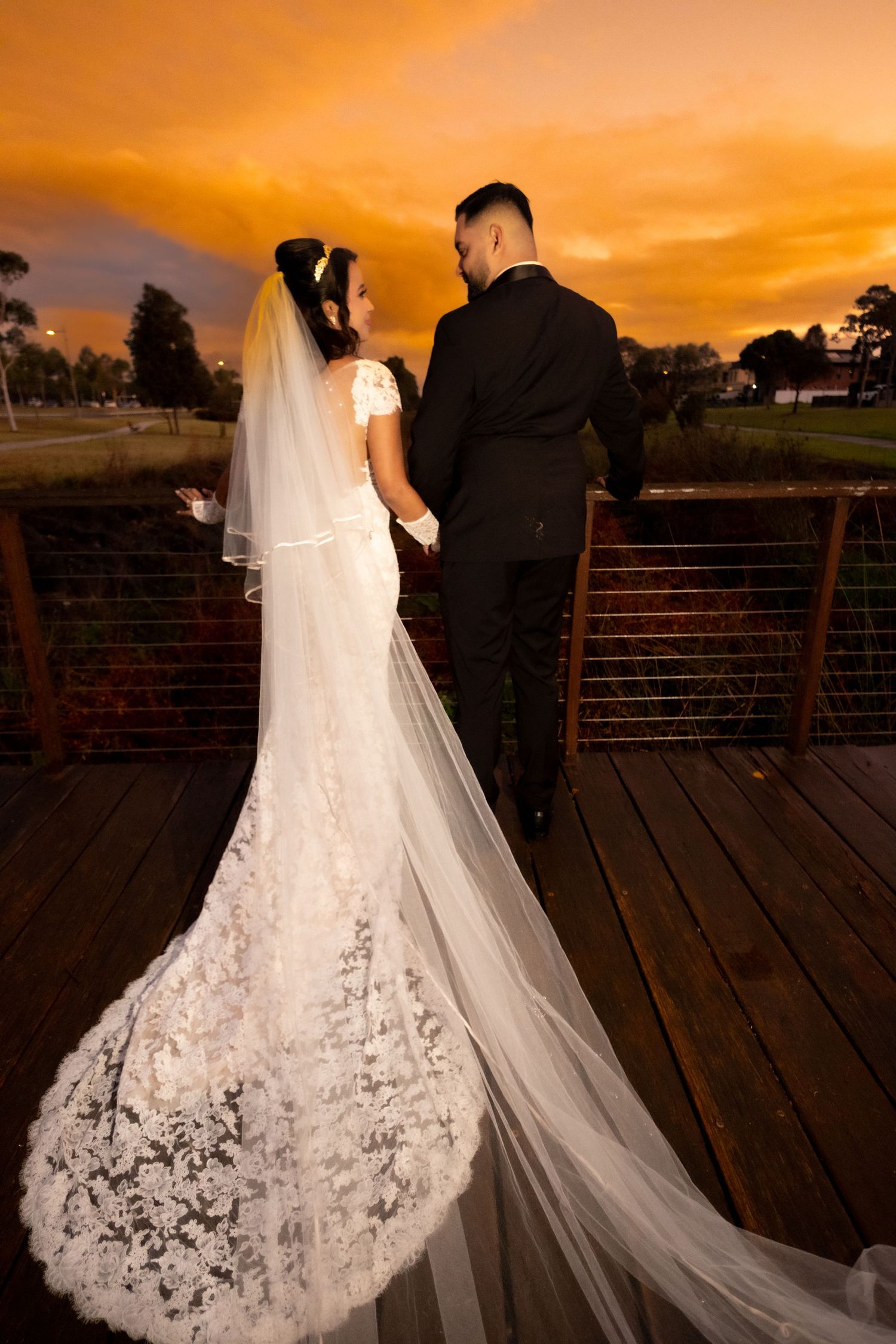 Bride and groom holding hands on wooden bridge at sunset, bride wearing lace gown with cathedral veil.