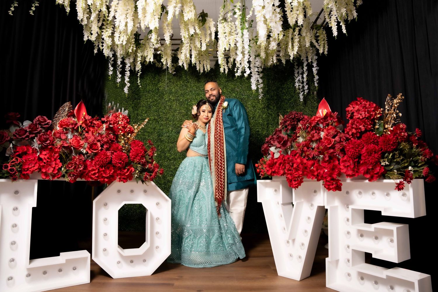 Couple poses in front of LOVE marquee letters with red floral backdrop.