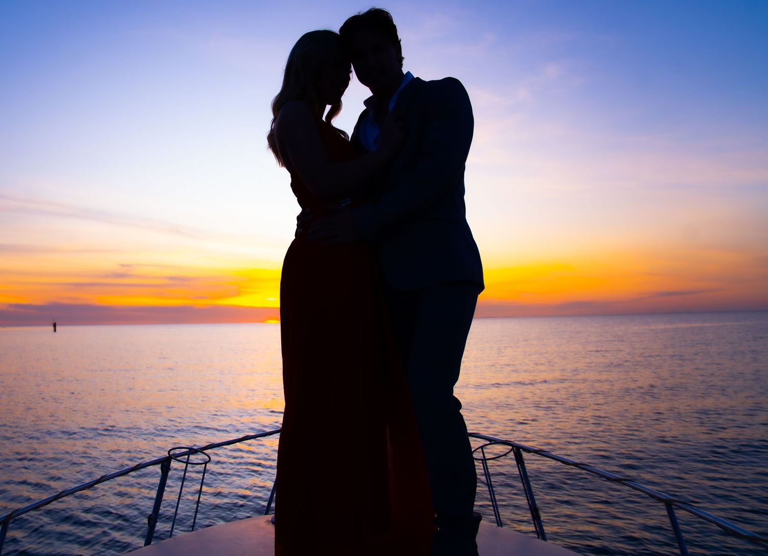 Romantic silhouette of couple embracing on boat deck against vibrant sunset over ocean.