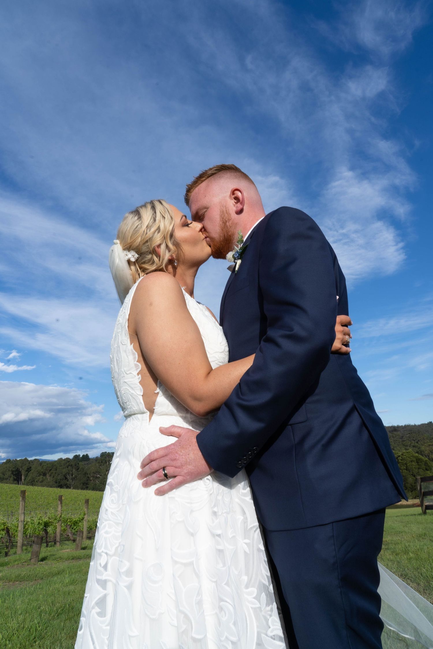 Bride and groom share a romantic kiss in vineyard setting under vivid blue sky.