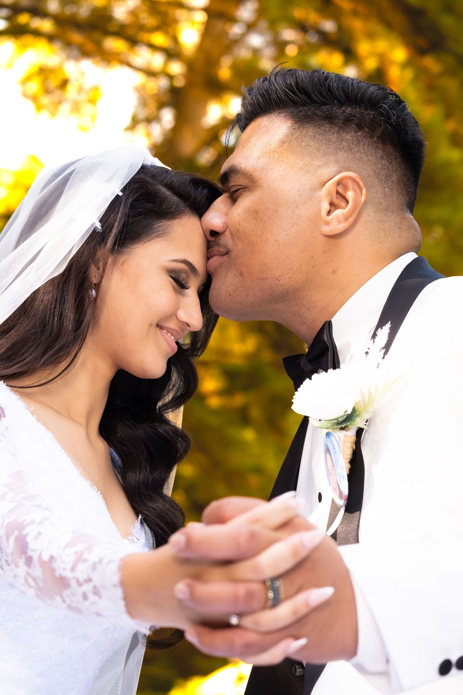 Groom tenderly kissing bride on forehead while holding hands in a golden autumn garden.