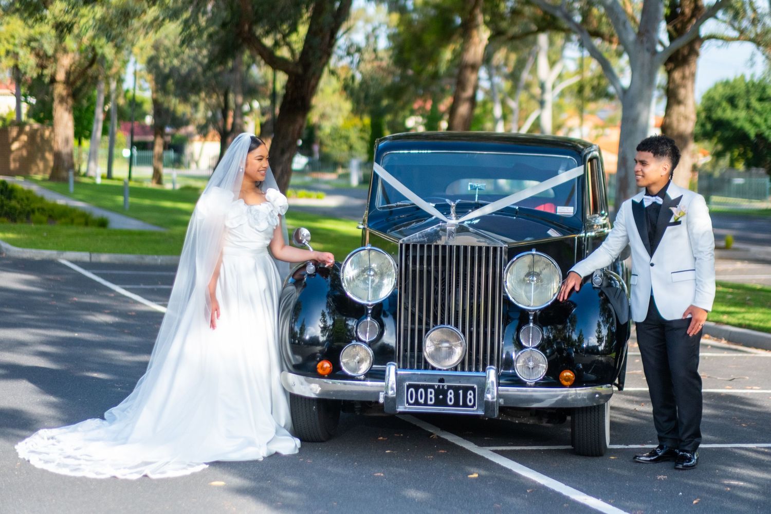 Bride and groom pose beside a vintage black Rolls-Royce in a tree-lined parking area.