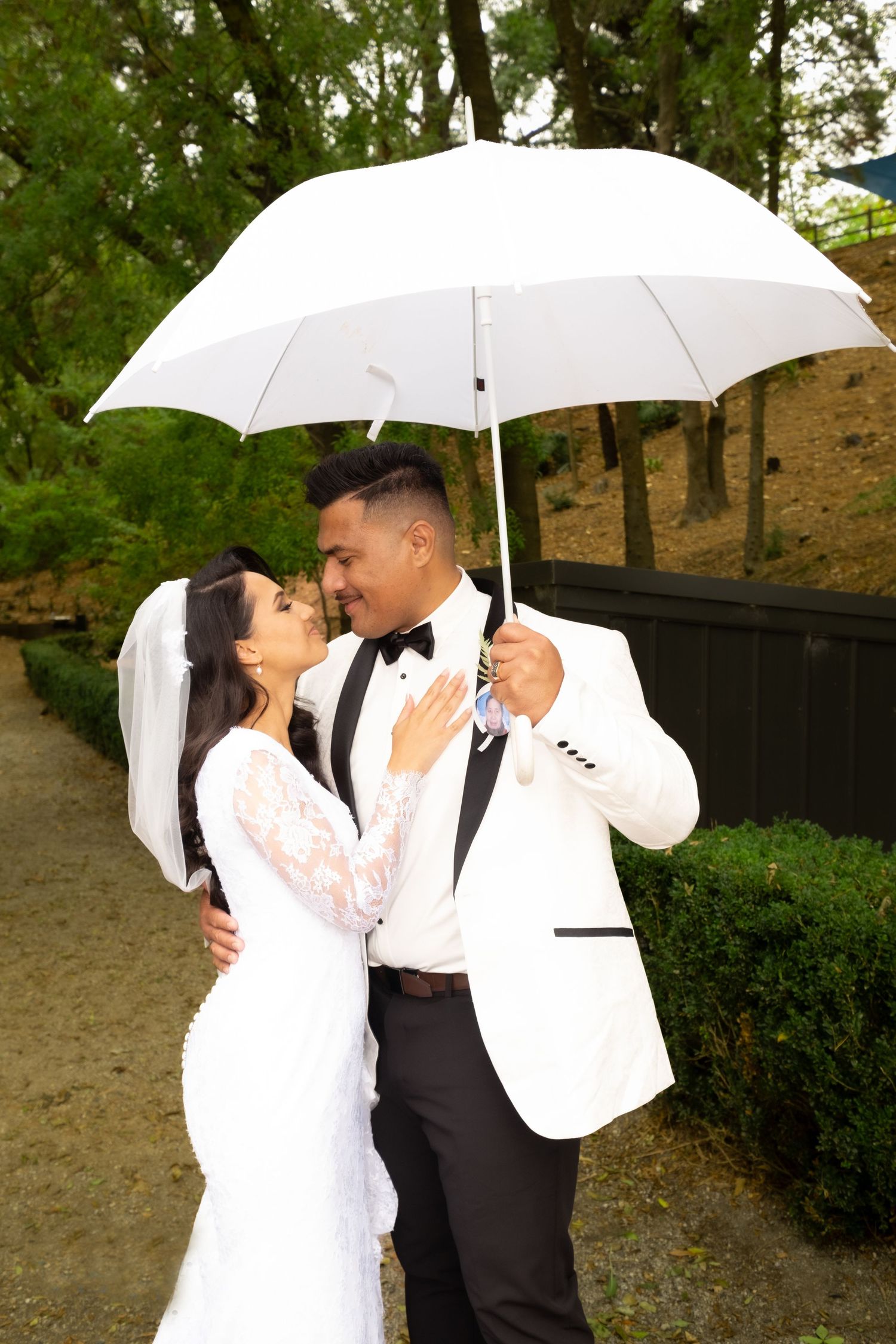 Bride and groom share romantic moment under white umbrella in lush garden.