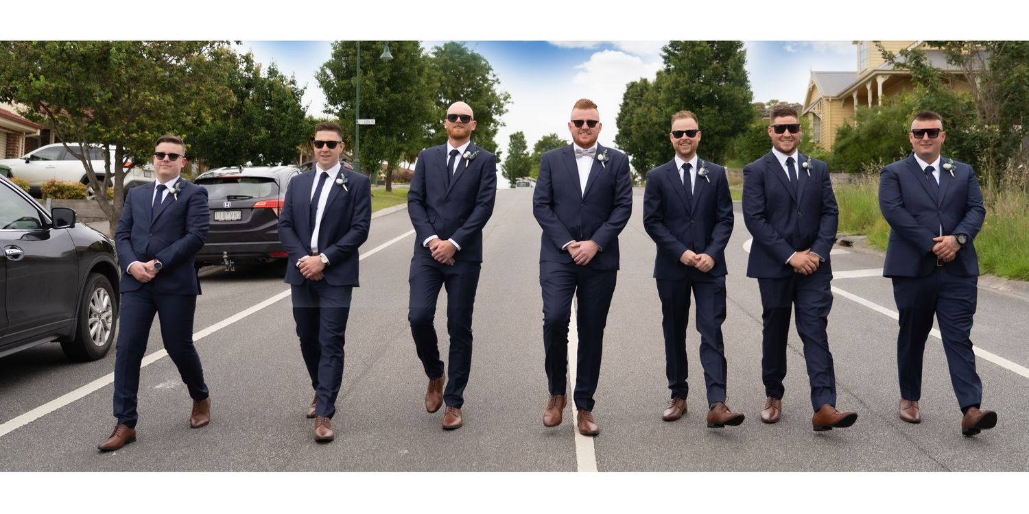 Seven groomsmen in matching navy suits and sunglasses posing confidently on a suburban street.
