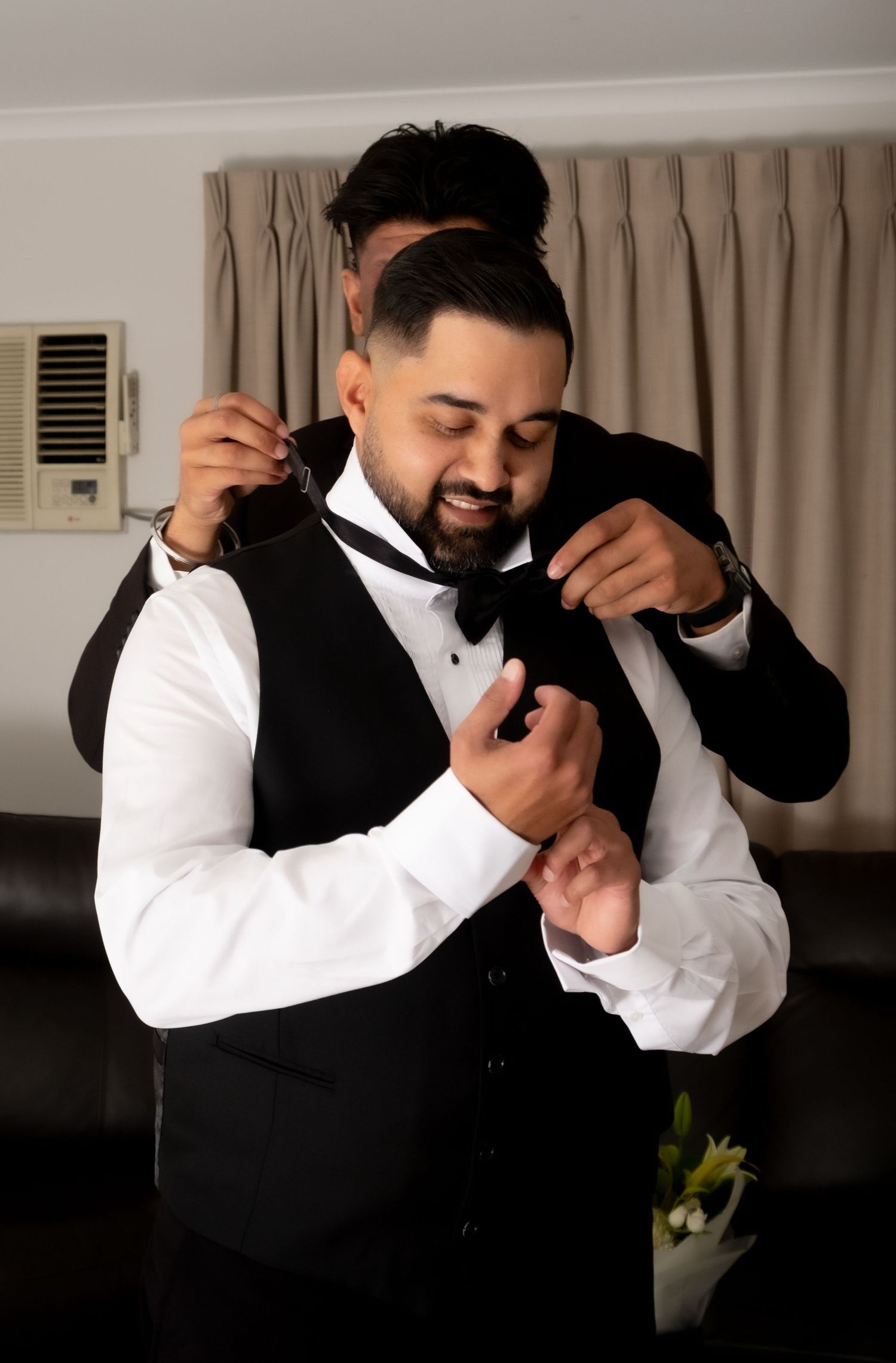 Groom in black tuxedo gets bow tie adjusted by groomsman while preparing for wedding ceremony.