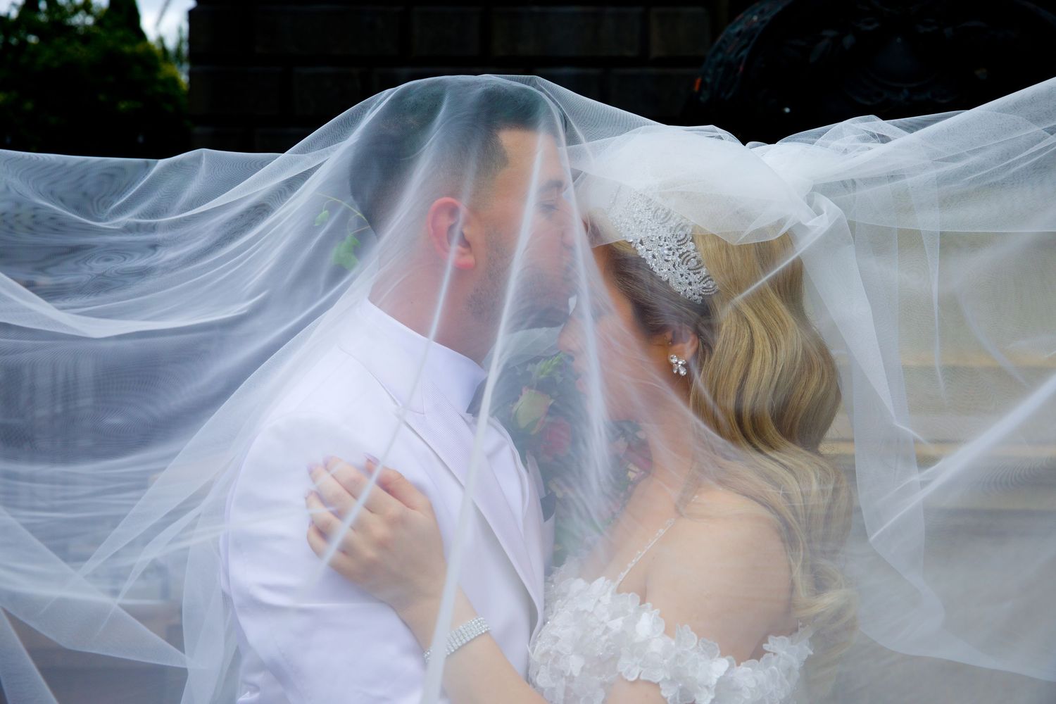 Bride and groom sharing romantic kiss beneath flowing white wedding veil outdoors.