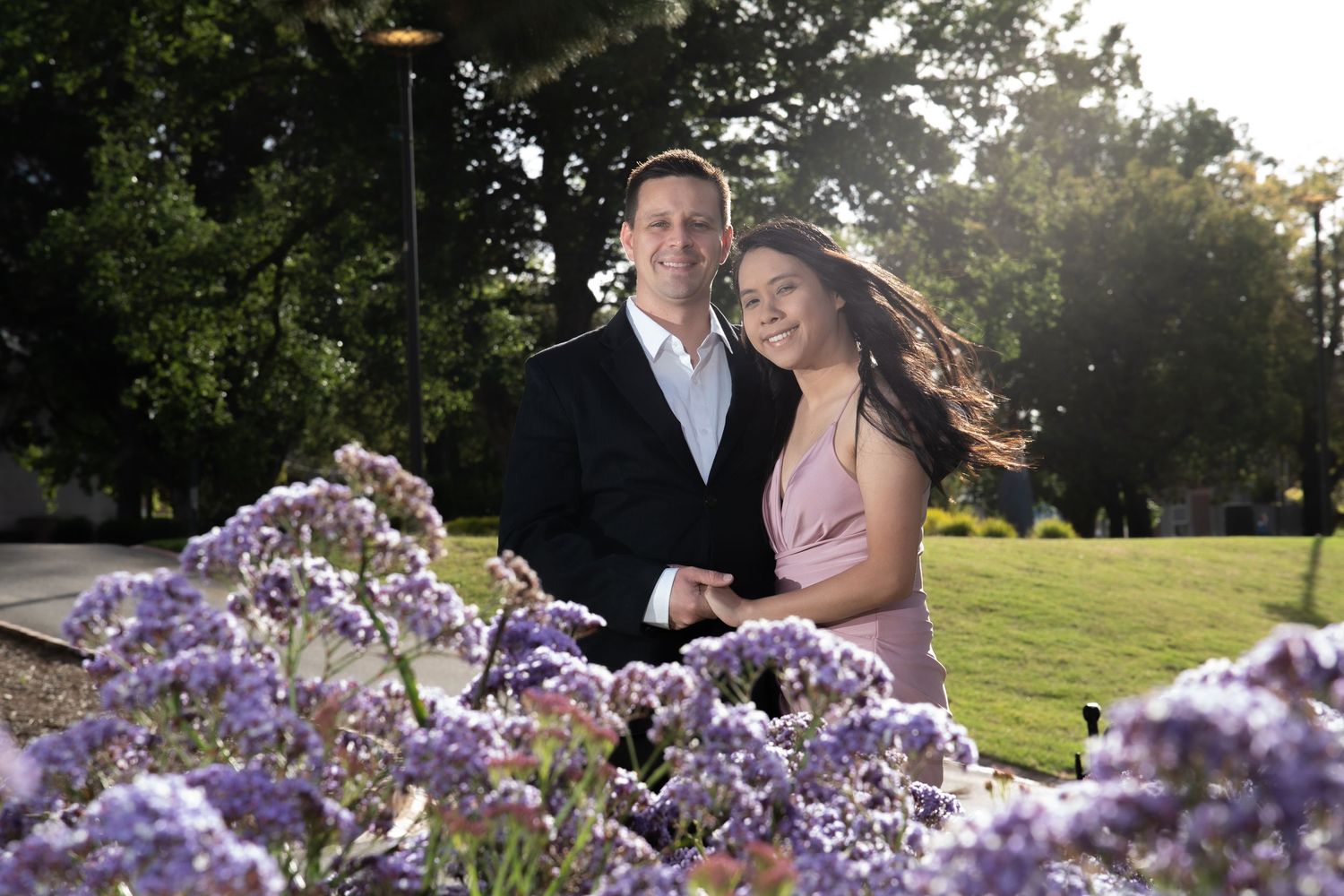 Couple posing among purple flowers at golden hour in a park, man in suit and woman in pink dress.