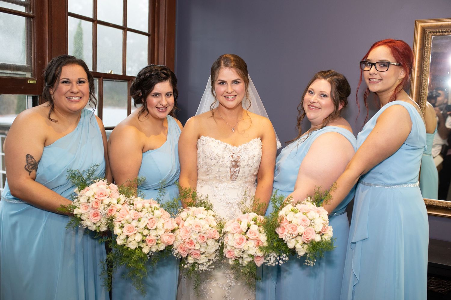 Bride in lace gown poses with four bridesmaids in blue dresses holding pink floral bouquets.