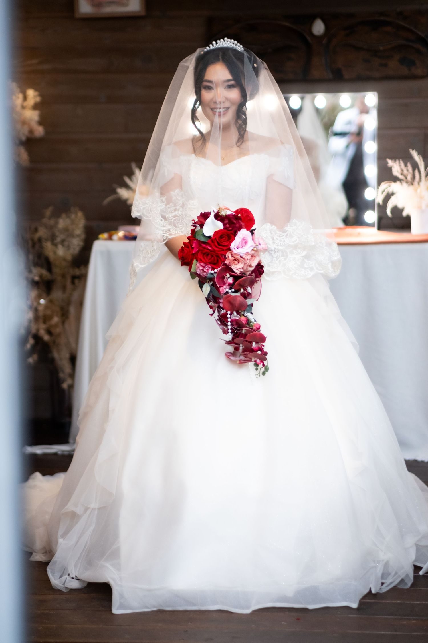 Bride in a white ball gown wedding dress with lace sleeves, veil, and tiara holding a red and pink cascading bouquet.