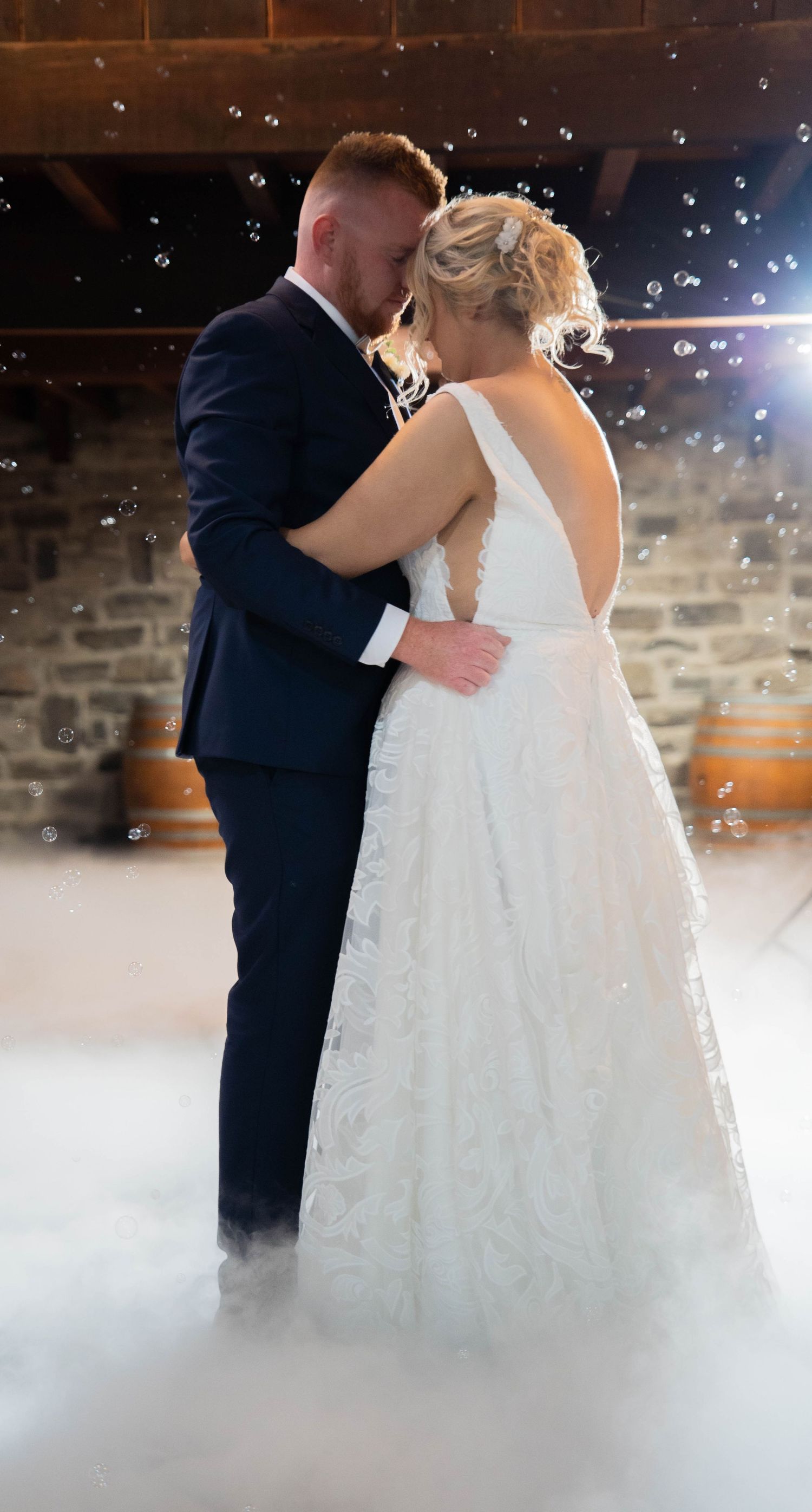 Bride and groom share their first dance surrounded by bubbles and atmospheric fog in a rustic stone barn.