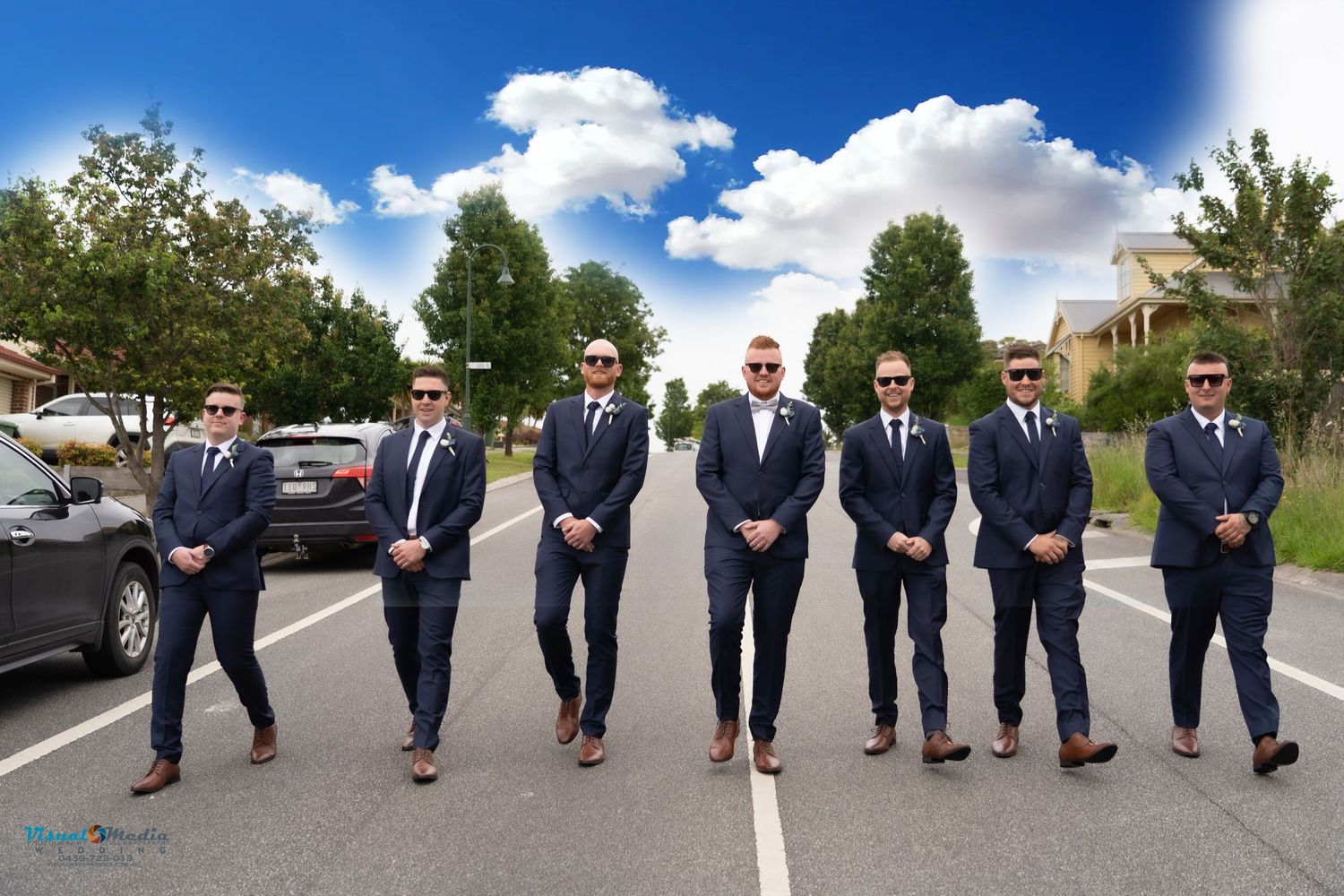 Seven groomsmen in matching navy suits and sunglasses pose confidently on a suburban road.