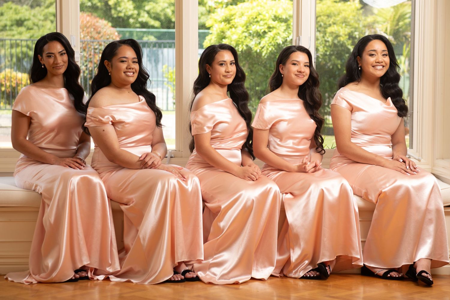 Five bridesmaids in blush satin off-shoulder gowns pose seated by a sunny window.