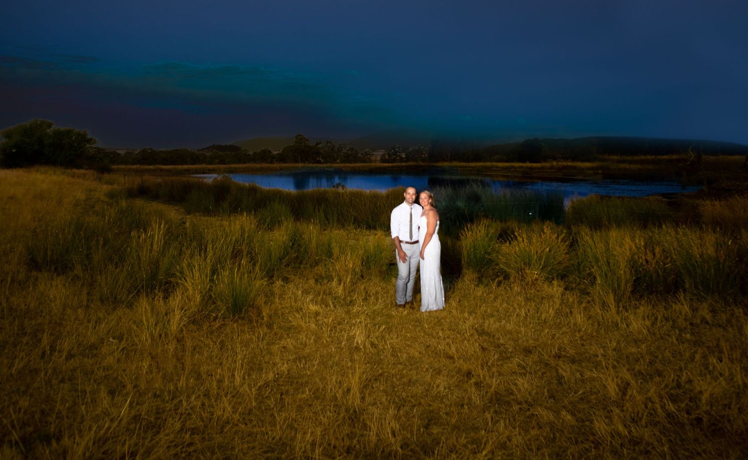 Bride and groom standing in golden grass field at dusk with a serene lake and dramatic blue sky backdrop.