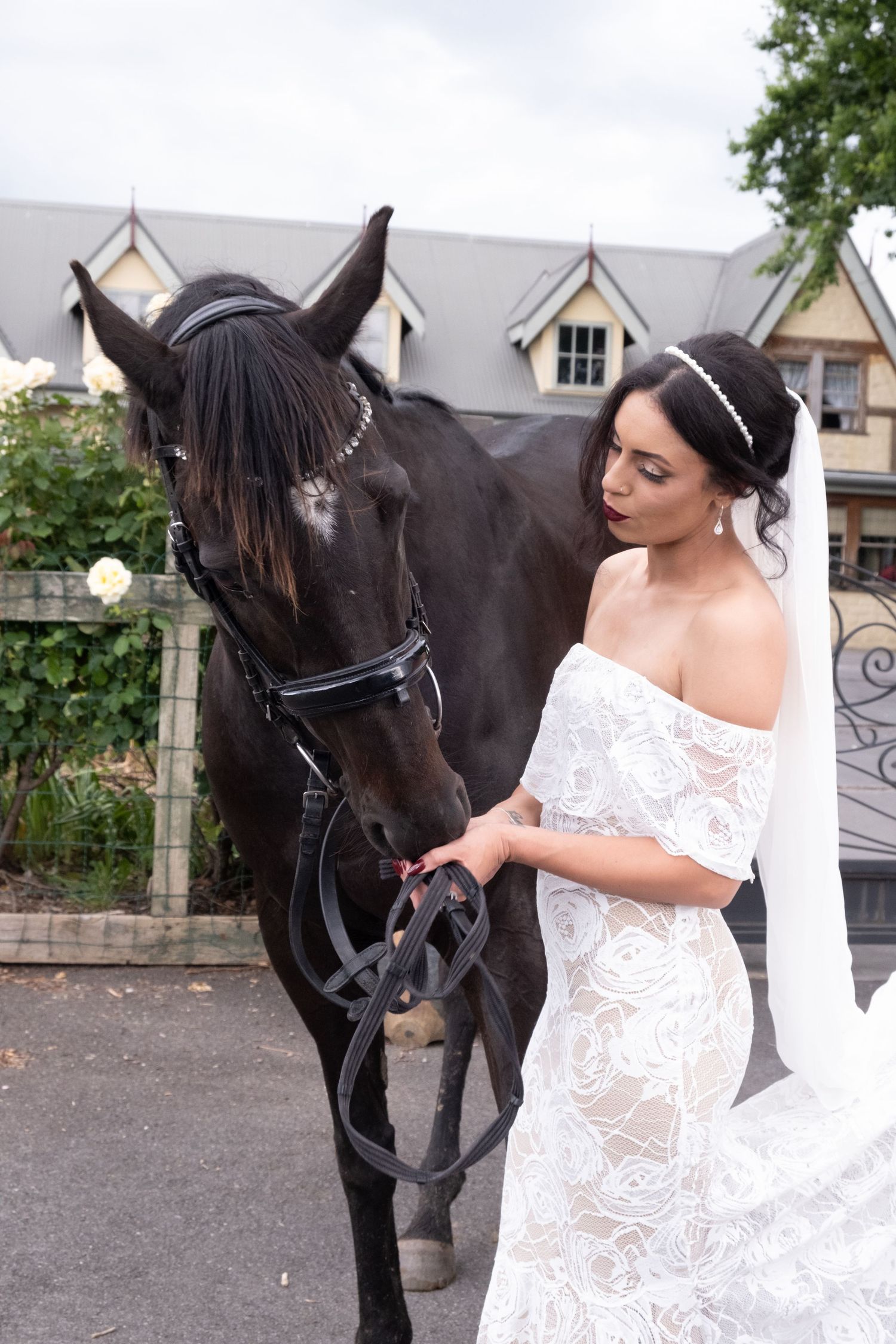 Bride in off-shoulder lace gown gently strokes a dark horse outside a stone building.