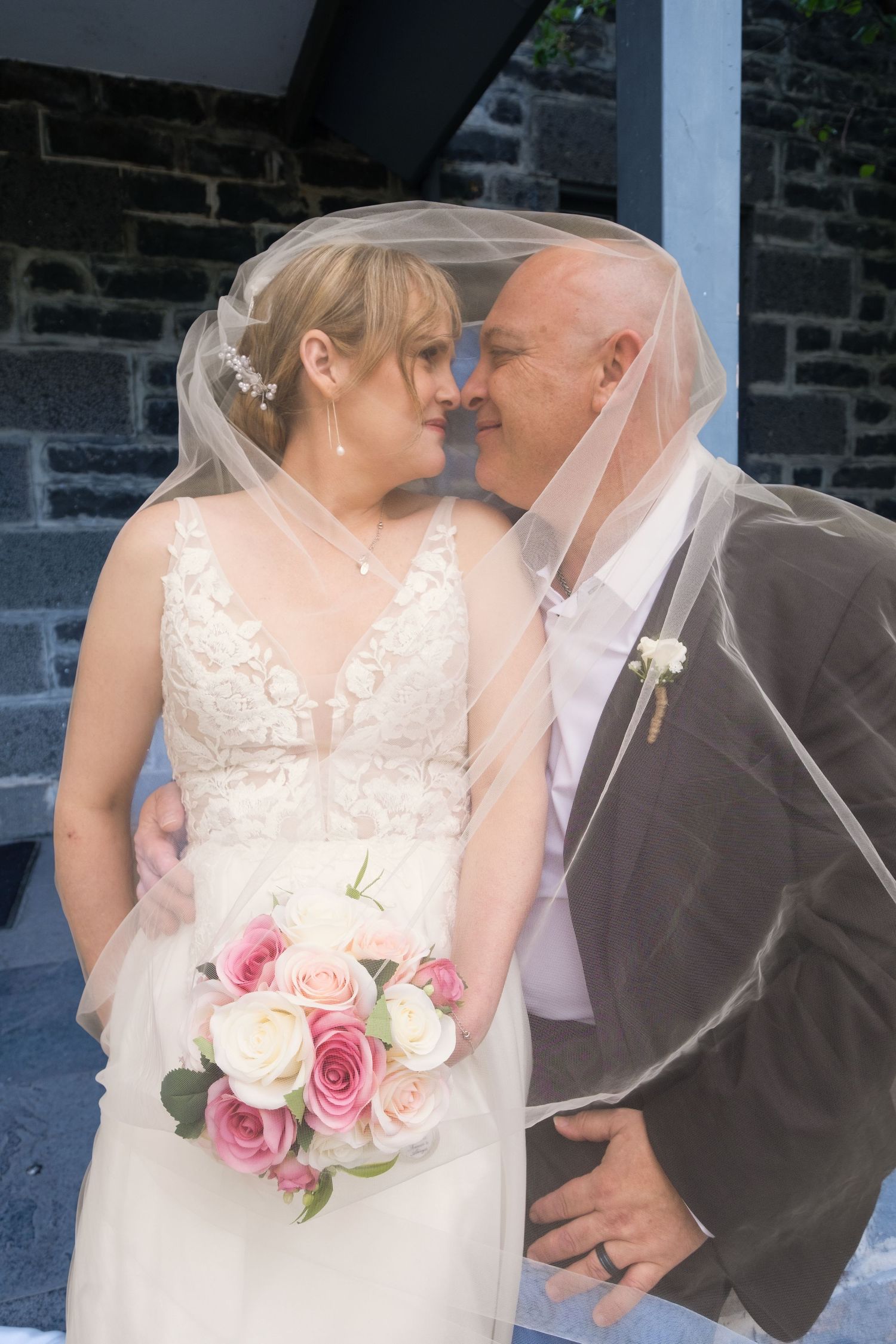 Bride and groom share a romantic moment under a wedding veil, with the bride holding a pink rose bouquet.