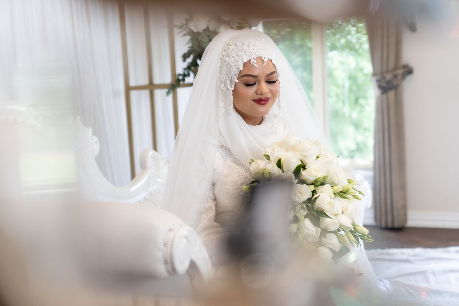 A bride in a white hijab and lace wedding dress holds a bouquet of white roses indoors.