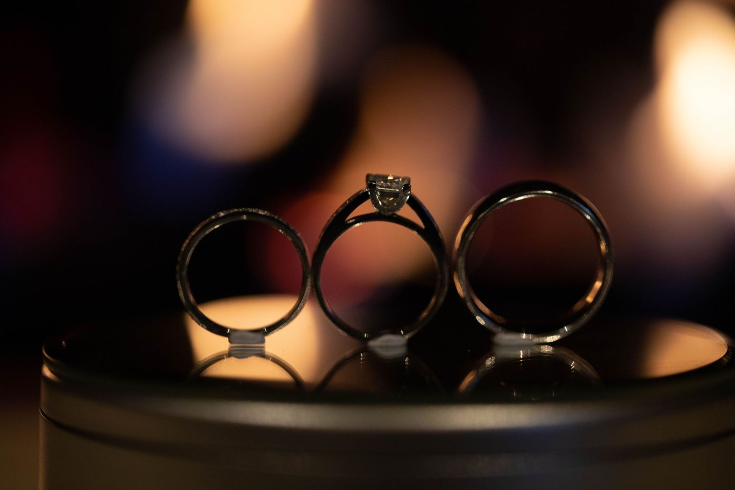 Three wedding rings, including a diamond solitaire engagement ring, displayed on a reflective surface with bokeh.