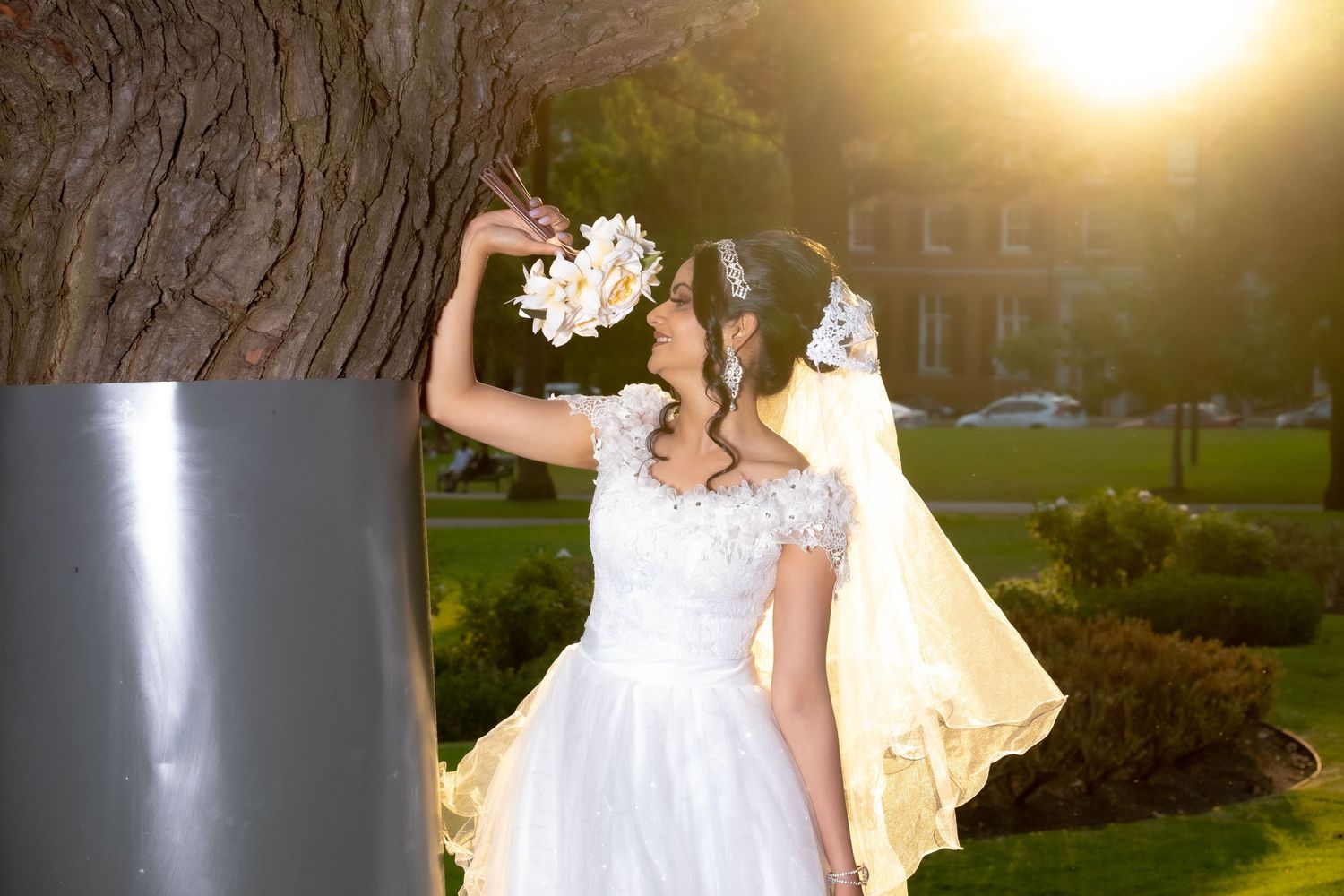 Bride in white lace gown with floral headpiece posing by a tree in golden sunset light.