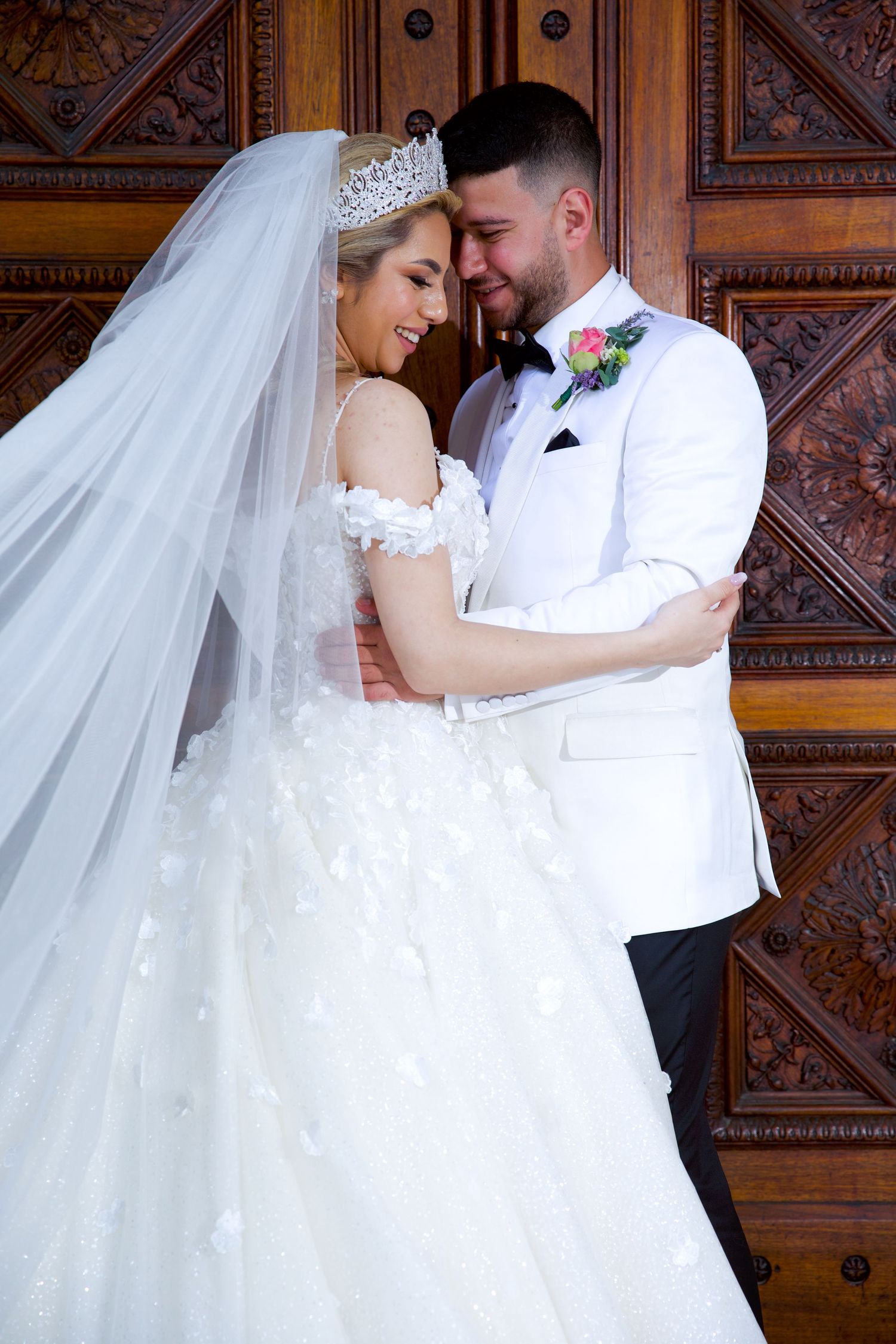 Smiling bride in crown and off-shoulder gown embraces groom in white tuxedo before ornate wooden doors.