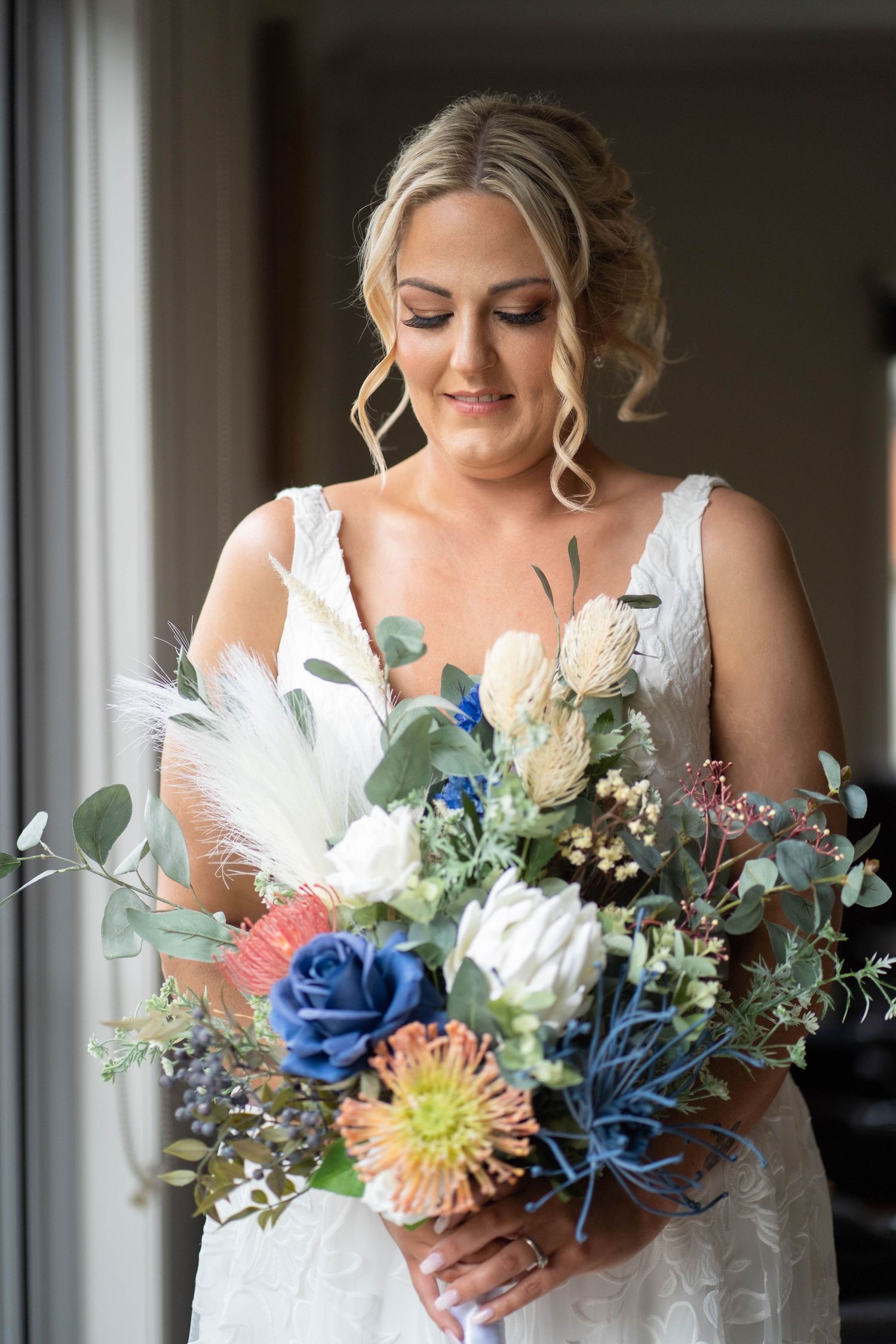 Elegant bride in lace gown admires her colorful boho wedding bouquet with proteas and roses.
