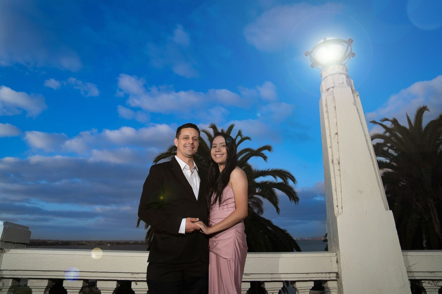 Couple posing together on a balcony under a dramatic blue sky with a glowing monument.