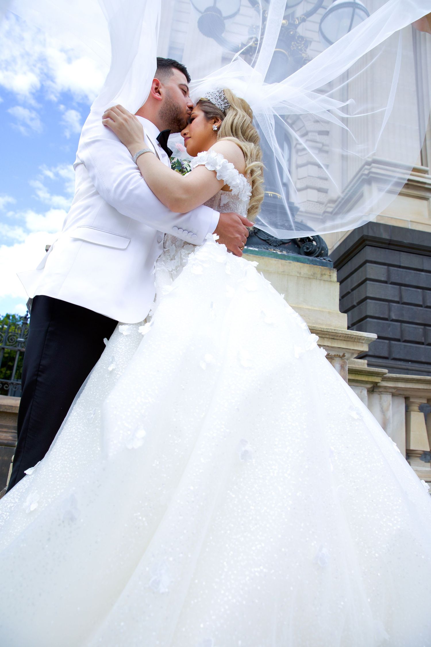 Bride and groom share a romantic kiss outdoors, her flowing white ballgown and veil billowing in the breeze.