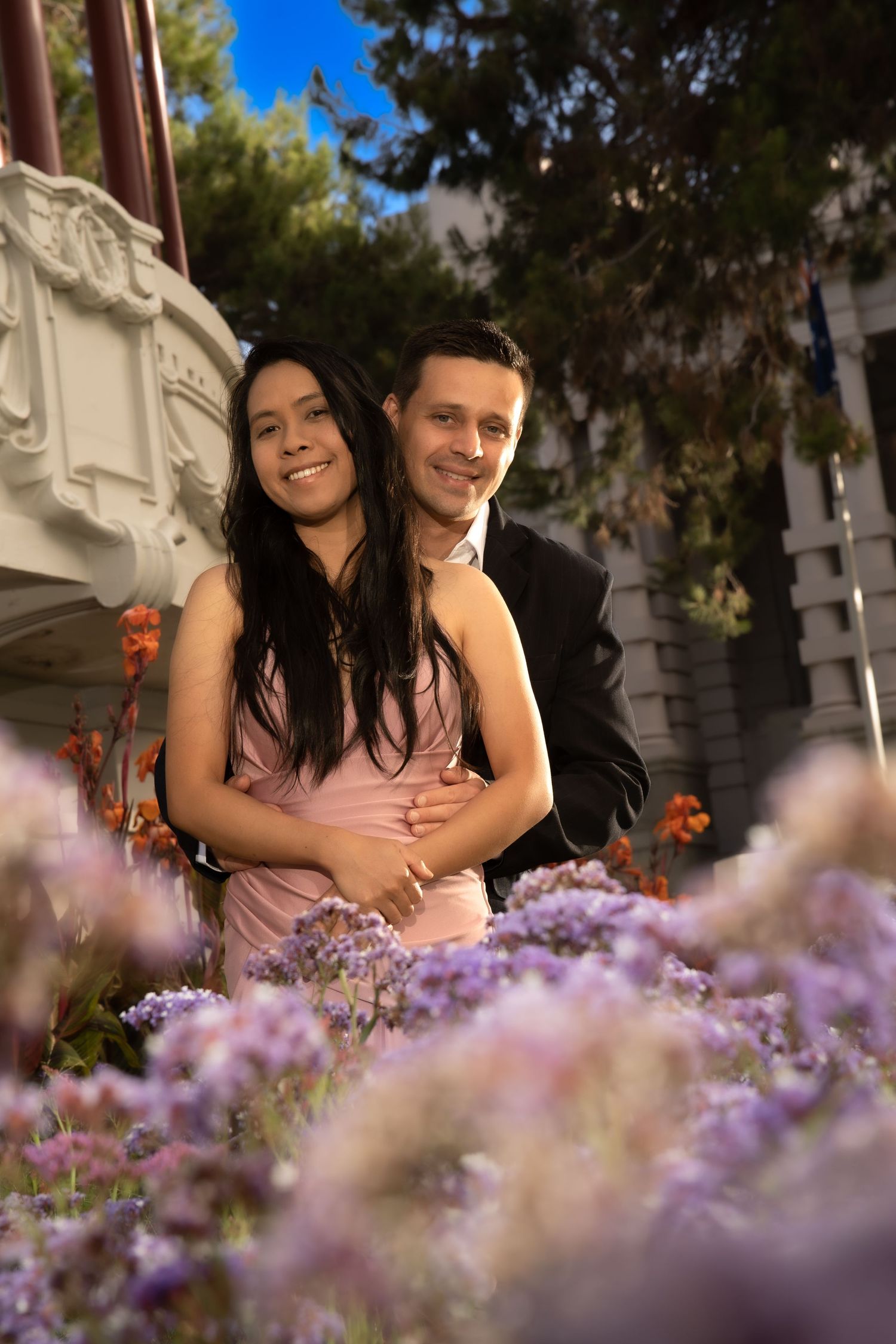 Couple embracing among purple flowers in a garden with historic architecture in the background.