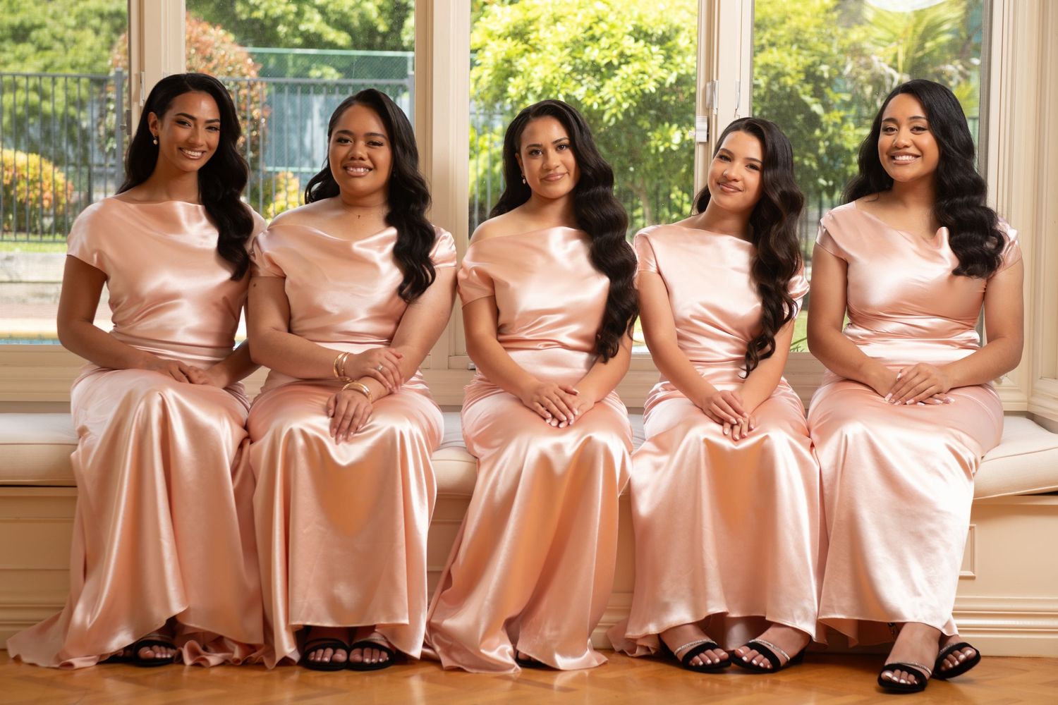 Five bridesmaids in matching rose gold satin dresses seated by a bright window smiling together.