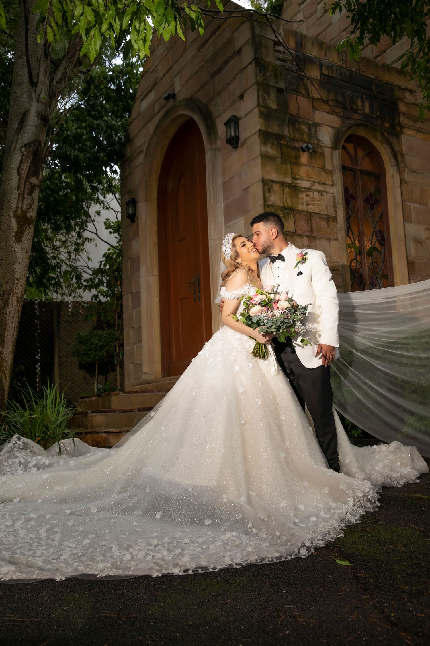 Bride in a floral ball gown and groom kissing before a stone chapel with a flowing veil.