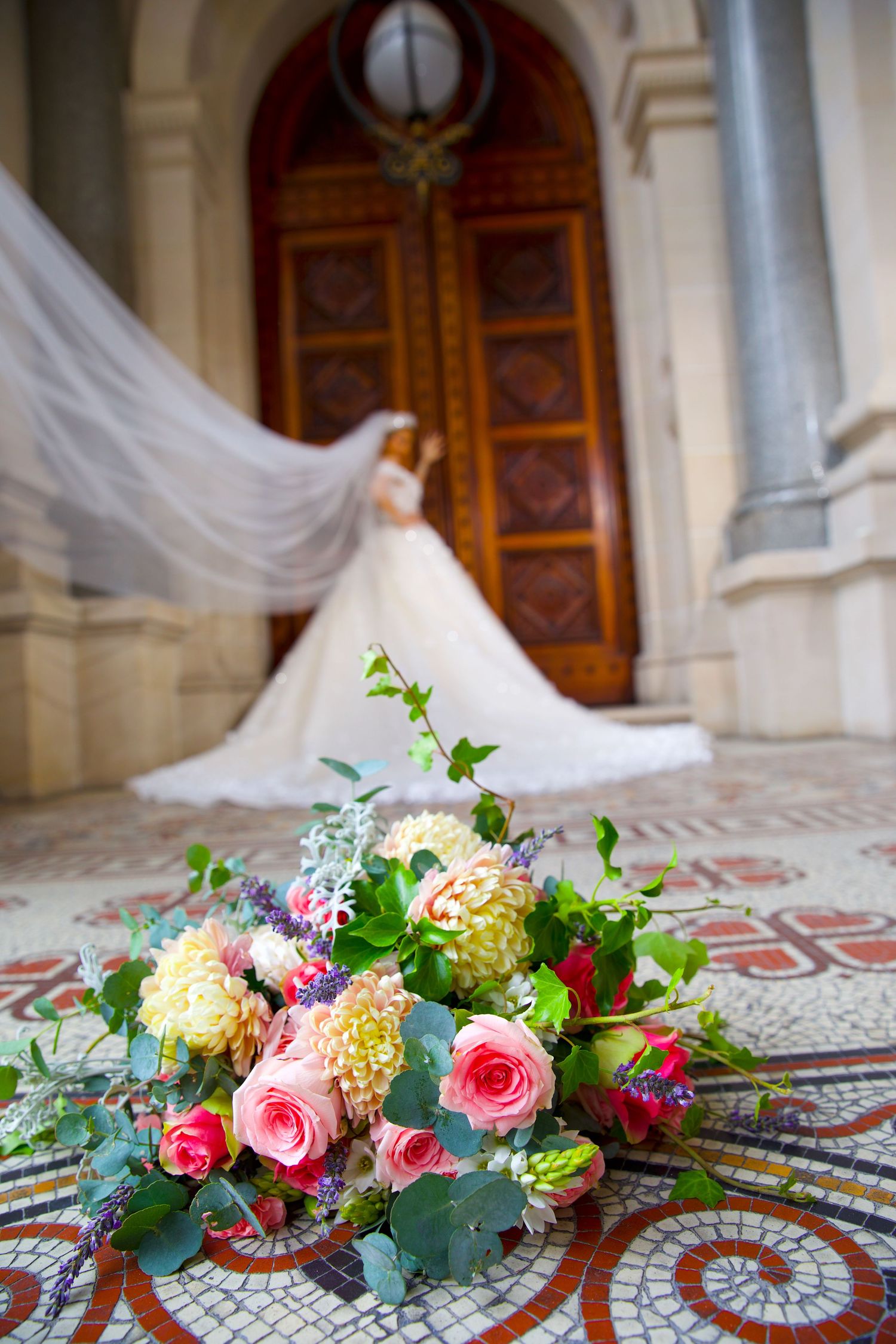 Colorful bridal bouquet of roses and dahlias in foreground with bride in flowing veil at church doors.