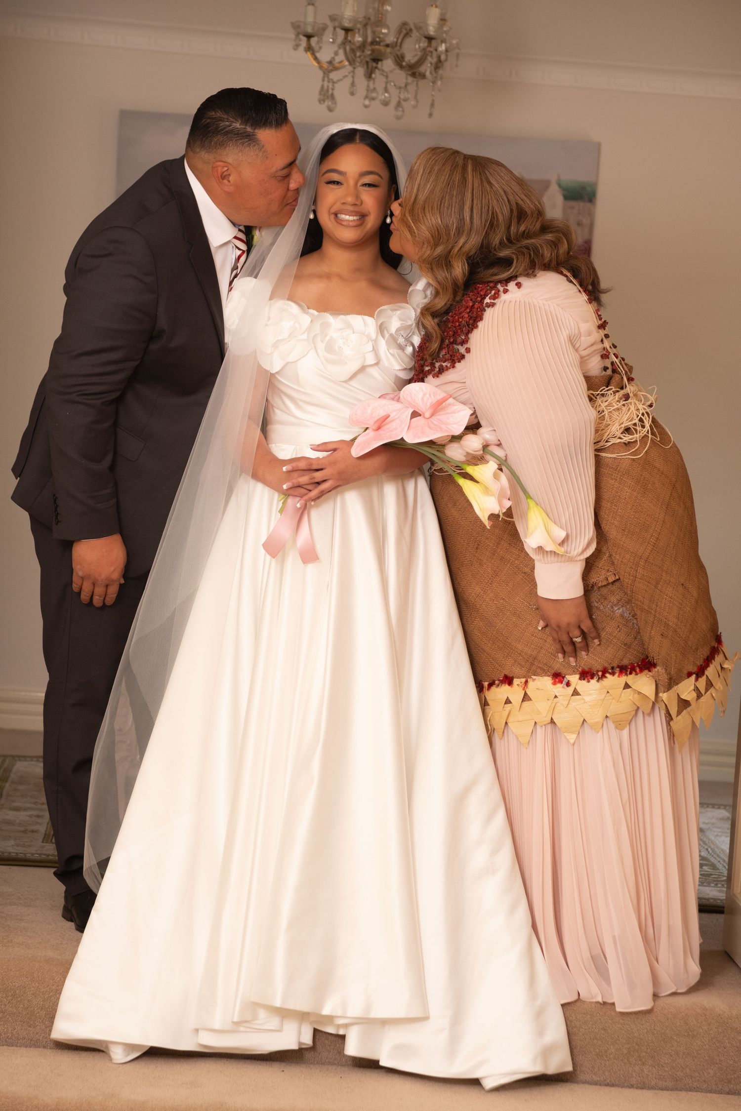 Bride in white gown receives kisses from parents in traditional and formal attire.