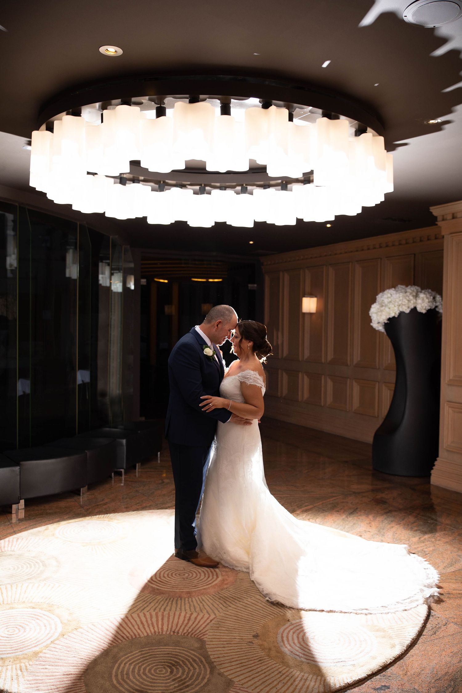 Bride and groom share romantic embrace under grand chandelier in luxurious hotel lobby.