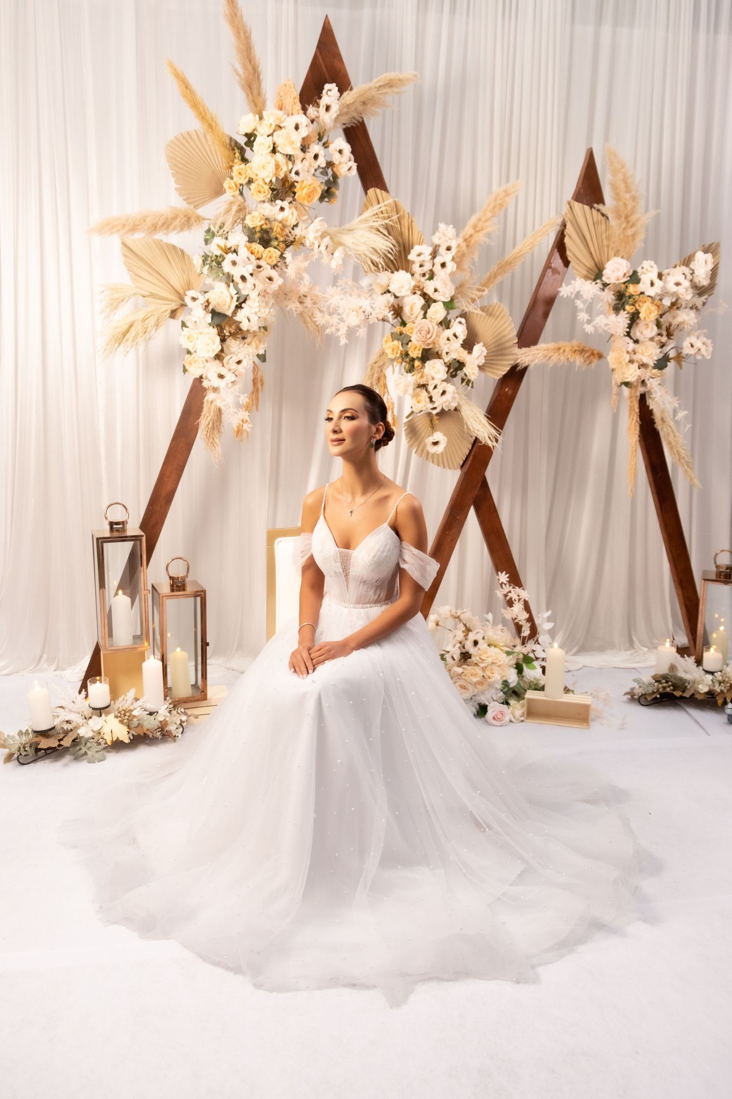 Bride in off-shoulder ball gown sitting before boho floral arch with dried pampas grass and white flowers.