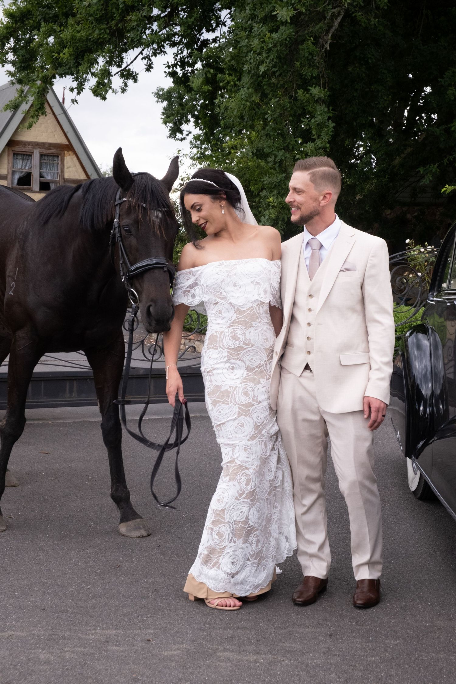 Bride in lace gown and groom in beige suit stand beside a black horse and vintage car.