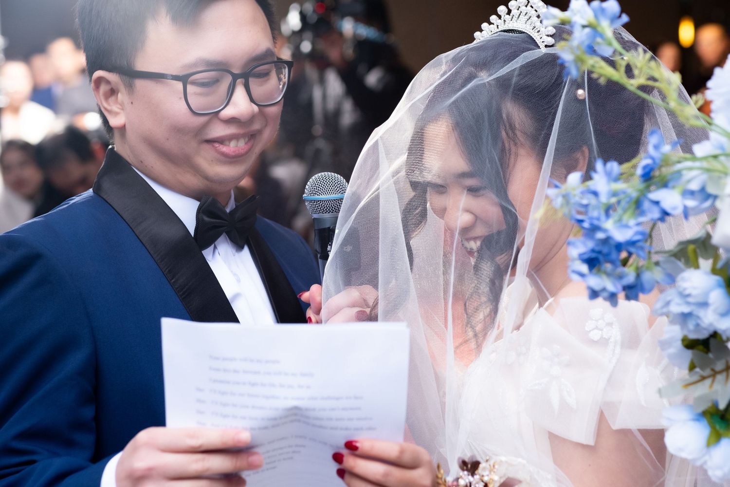 Groom in tuxedo and bride in veil reading wedding vows together at a romantic ceremony with blue floral decor.