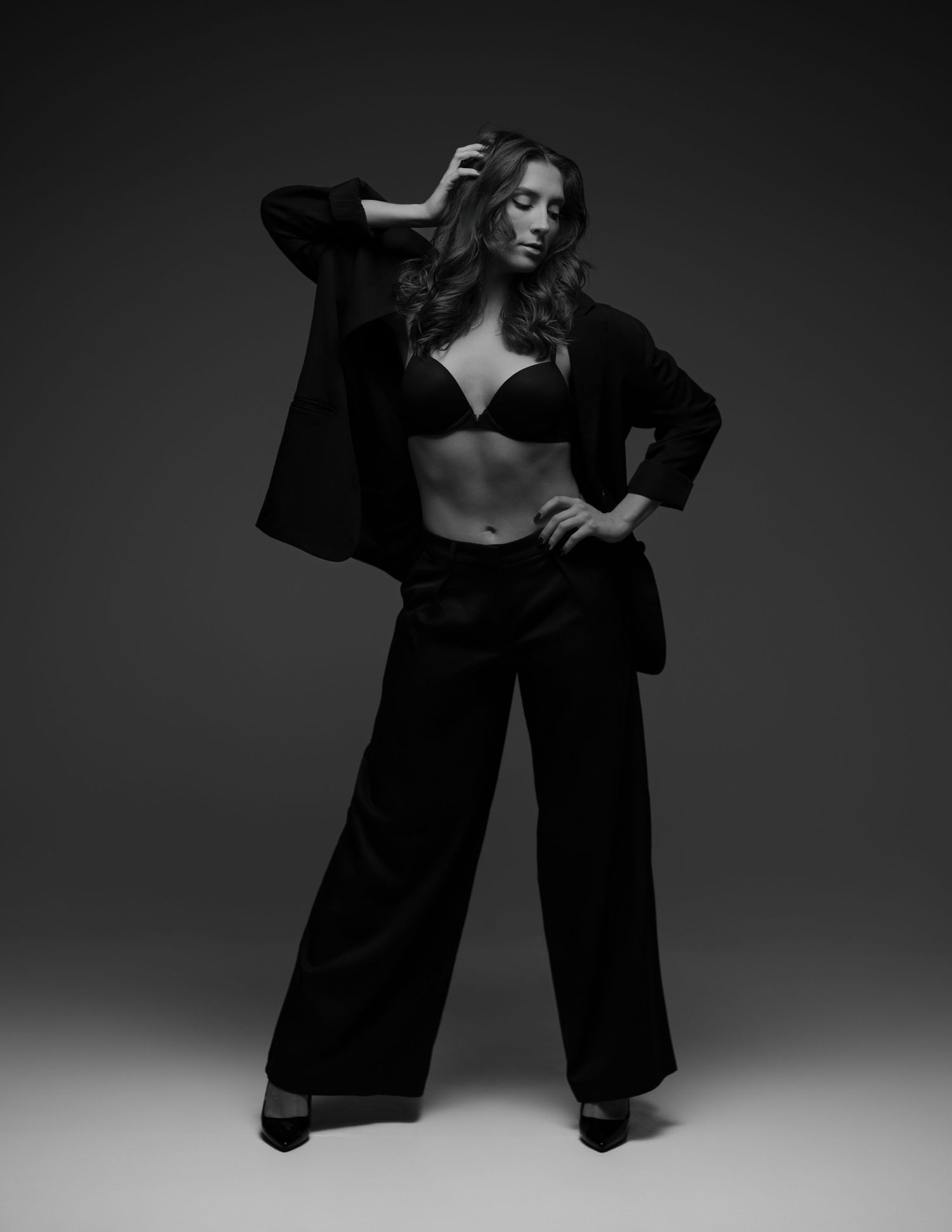 Black and white fashion photo of a woman in wide-leg trousers, blazer, and bralette posing confidently in studio.