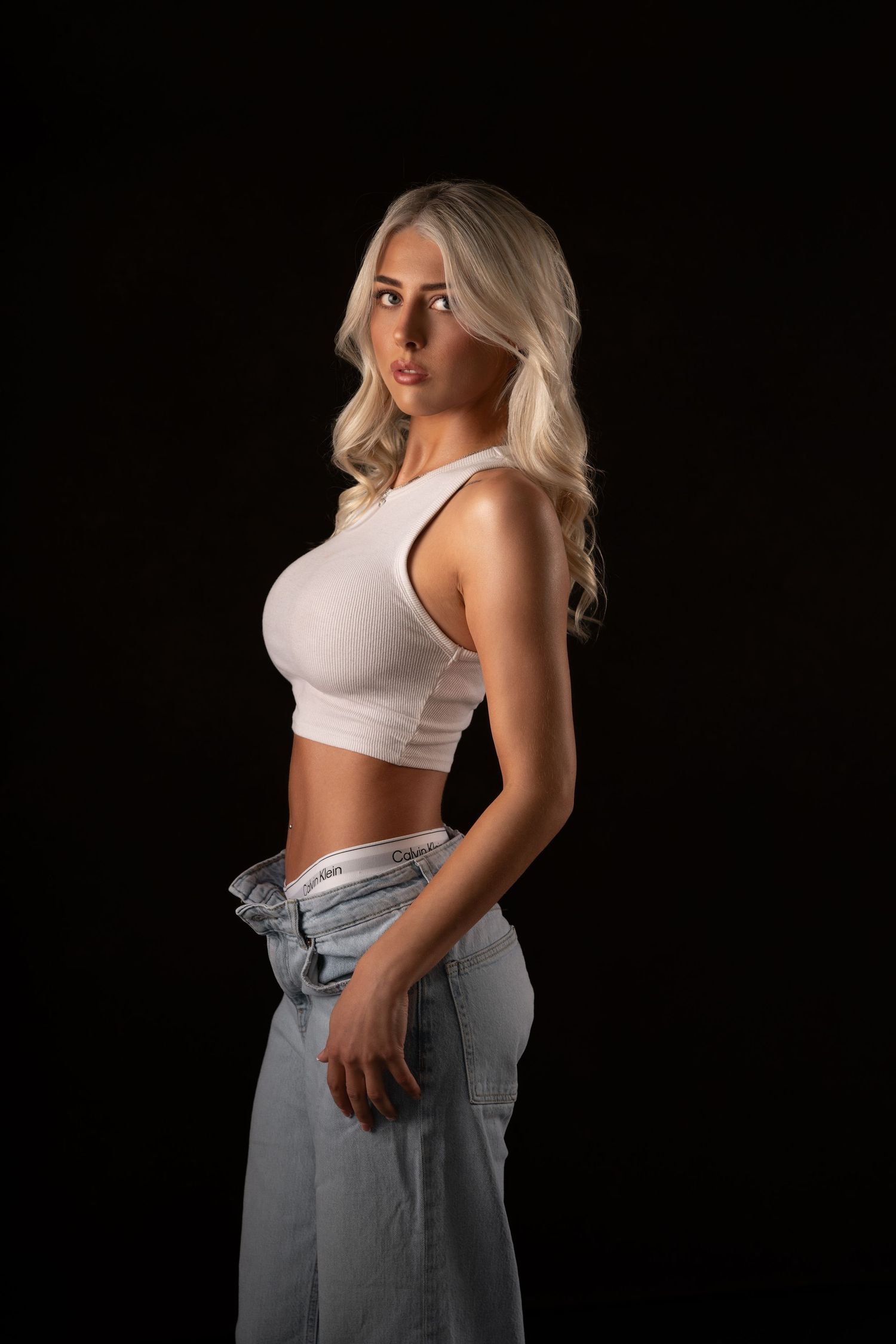 Blonde woman in white crop top and light wash jeans posing against black background in studio portrait.