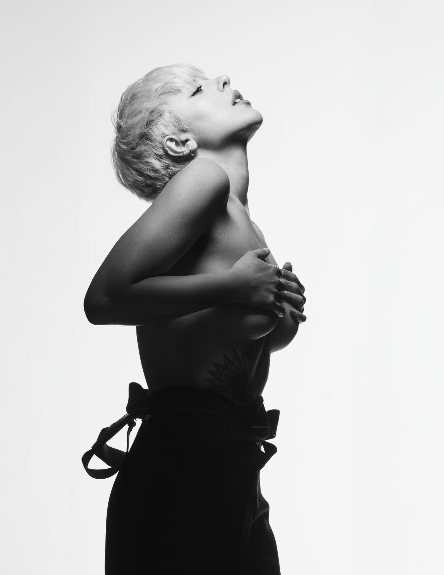 Black and white portrait of a woman with short blonde hair, posing topless with arms crossed over chest.