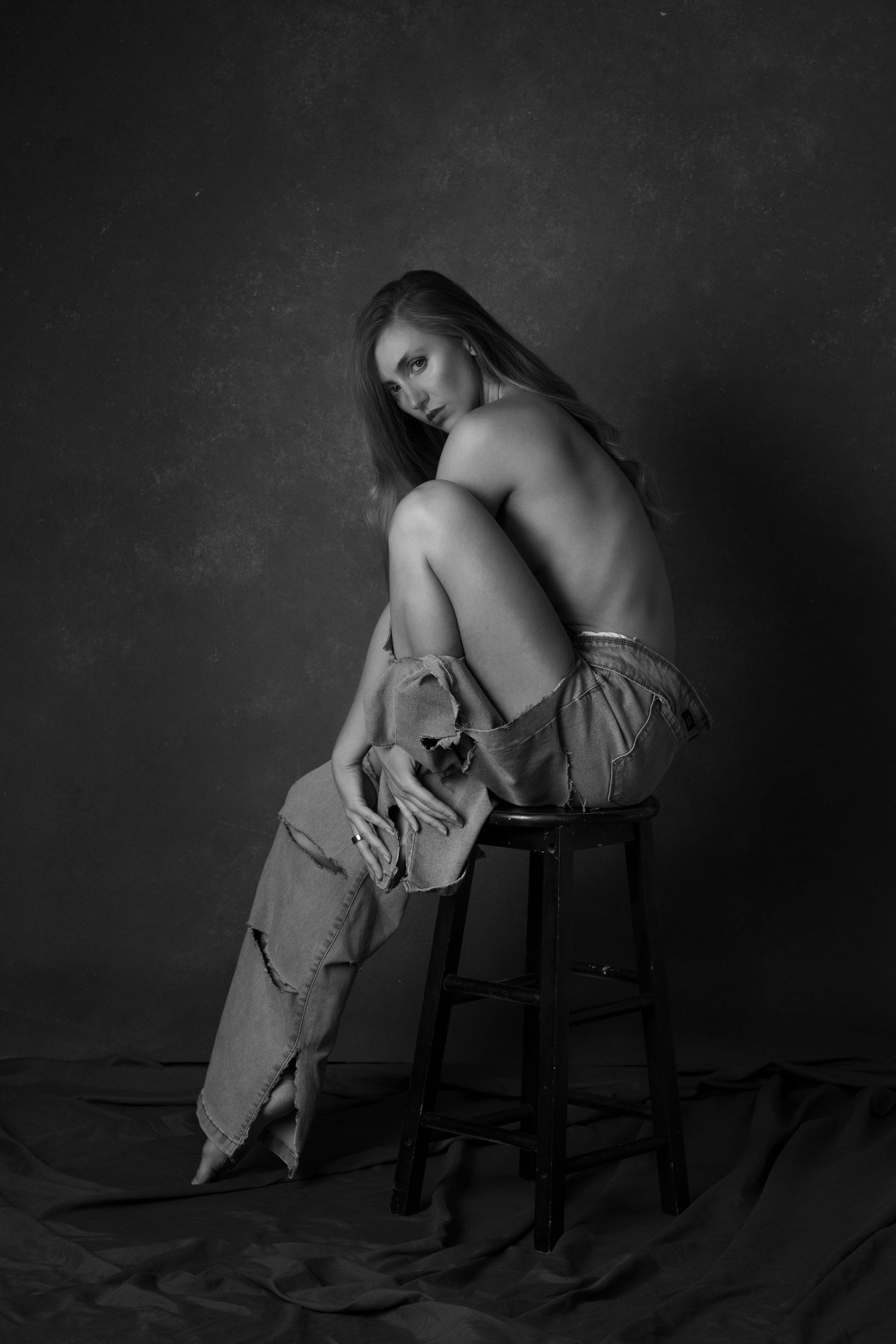 Black and white fine art photo of a young woman seated on a stool, posed gracefully with draped fabric.