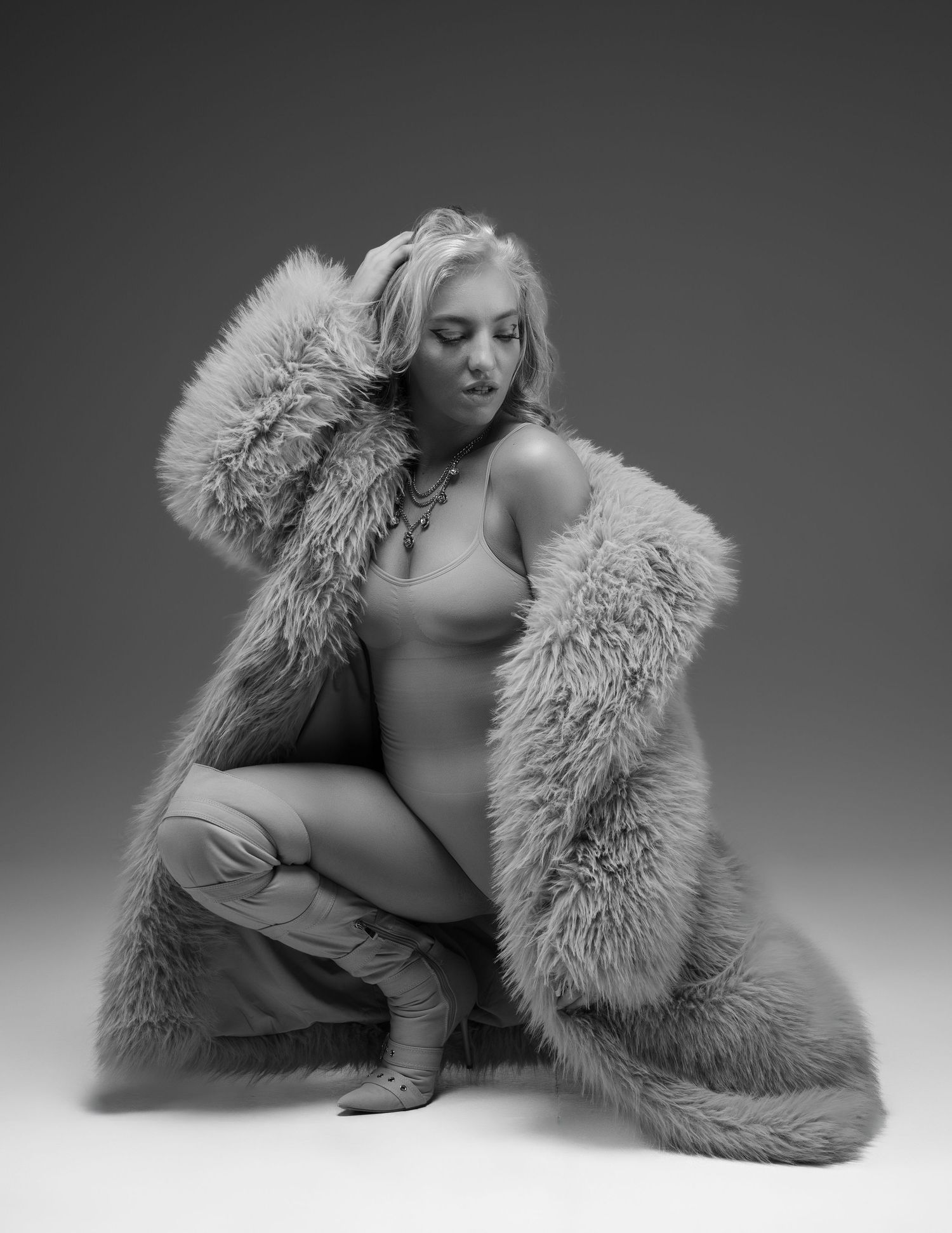 Black and white photo of a blonde woman posing in a oversized faux fur coat and boots in a studio.