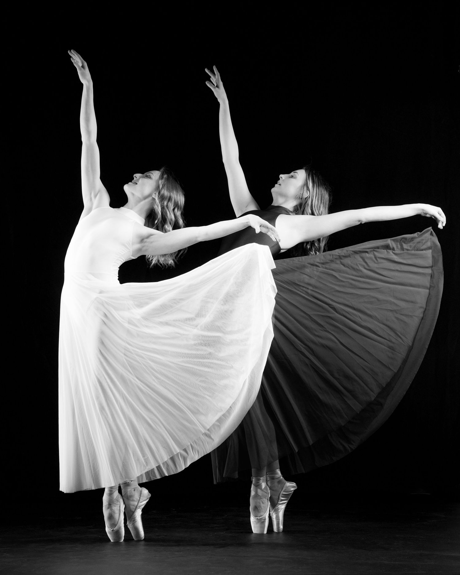 Two ballet dancers in white and dark gowns perform an elegant arabesque pose against a black background.