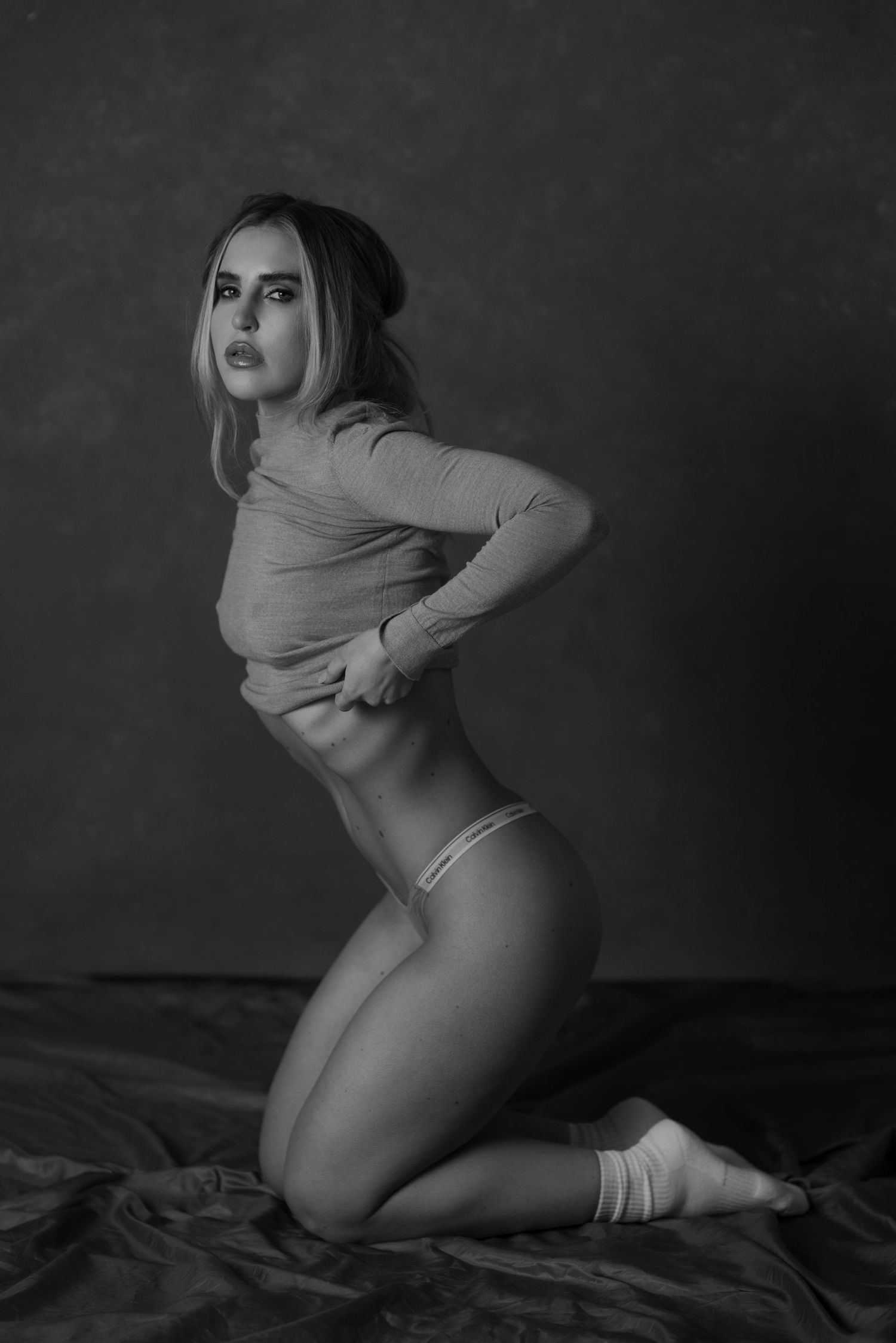 Black and white fine art photo of a woman kneeling in crop top and underwear against dark studio background.