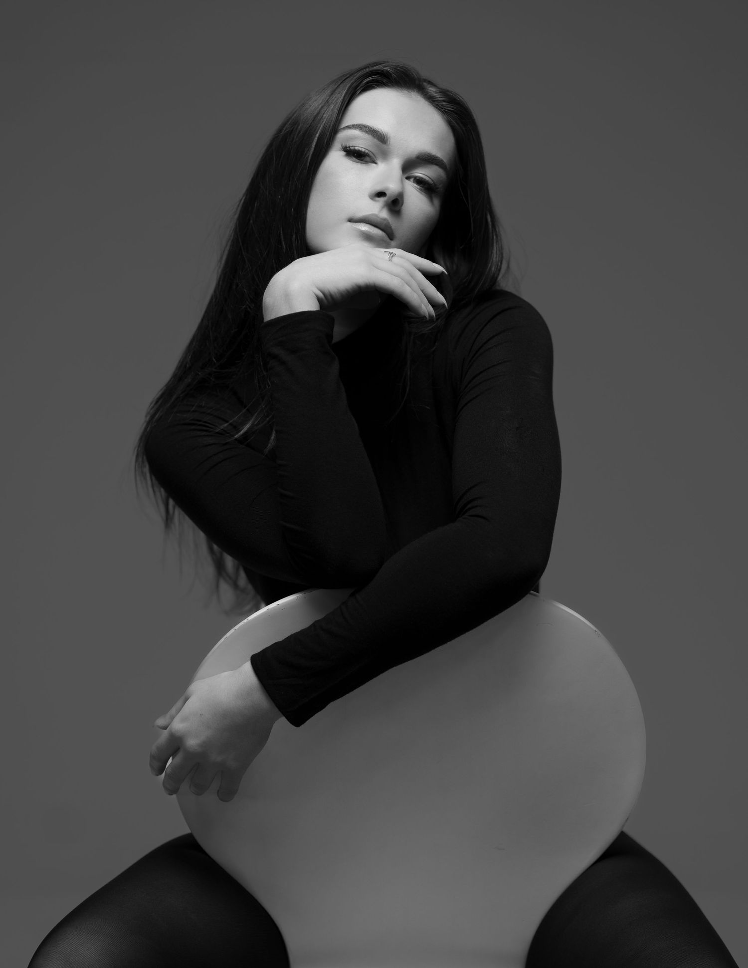 Black and white portrait of a young woman in a black turtleneck, posing elegantly on a modern white chair.