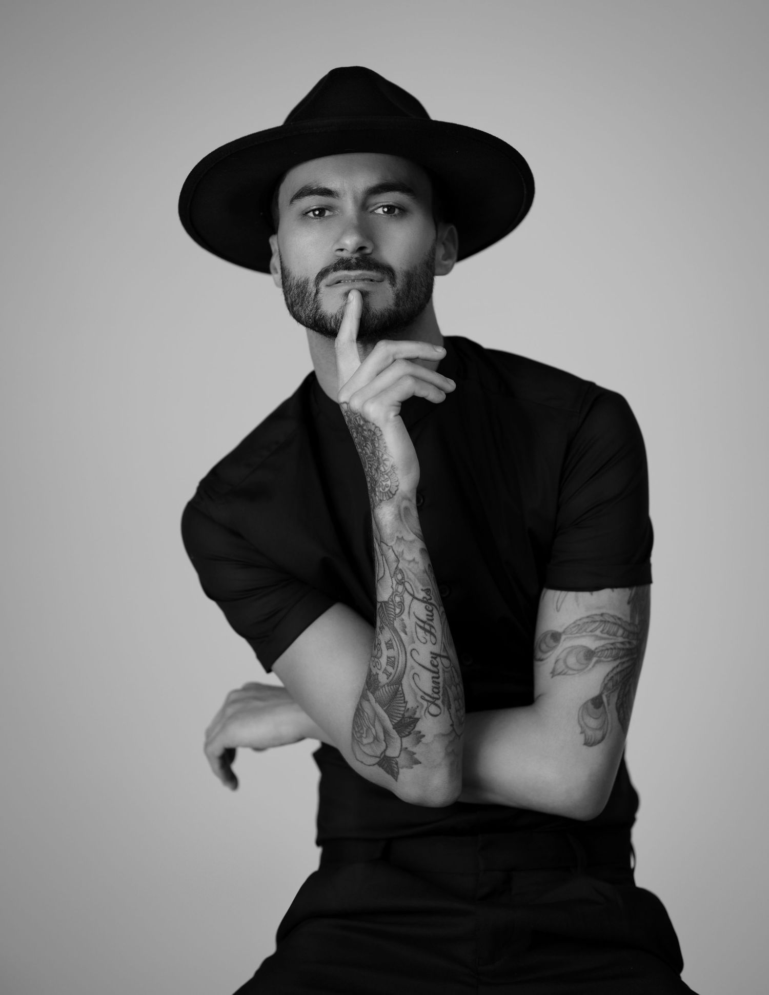 Black and white portrait of tattooed man in black hat and shirt posing thoughtfully with finger on chin.