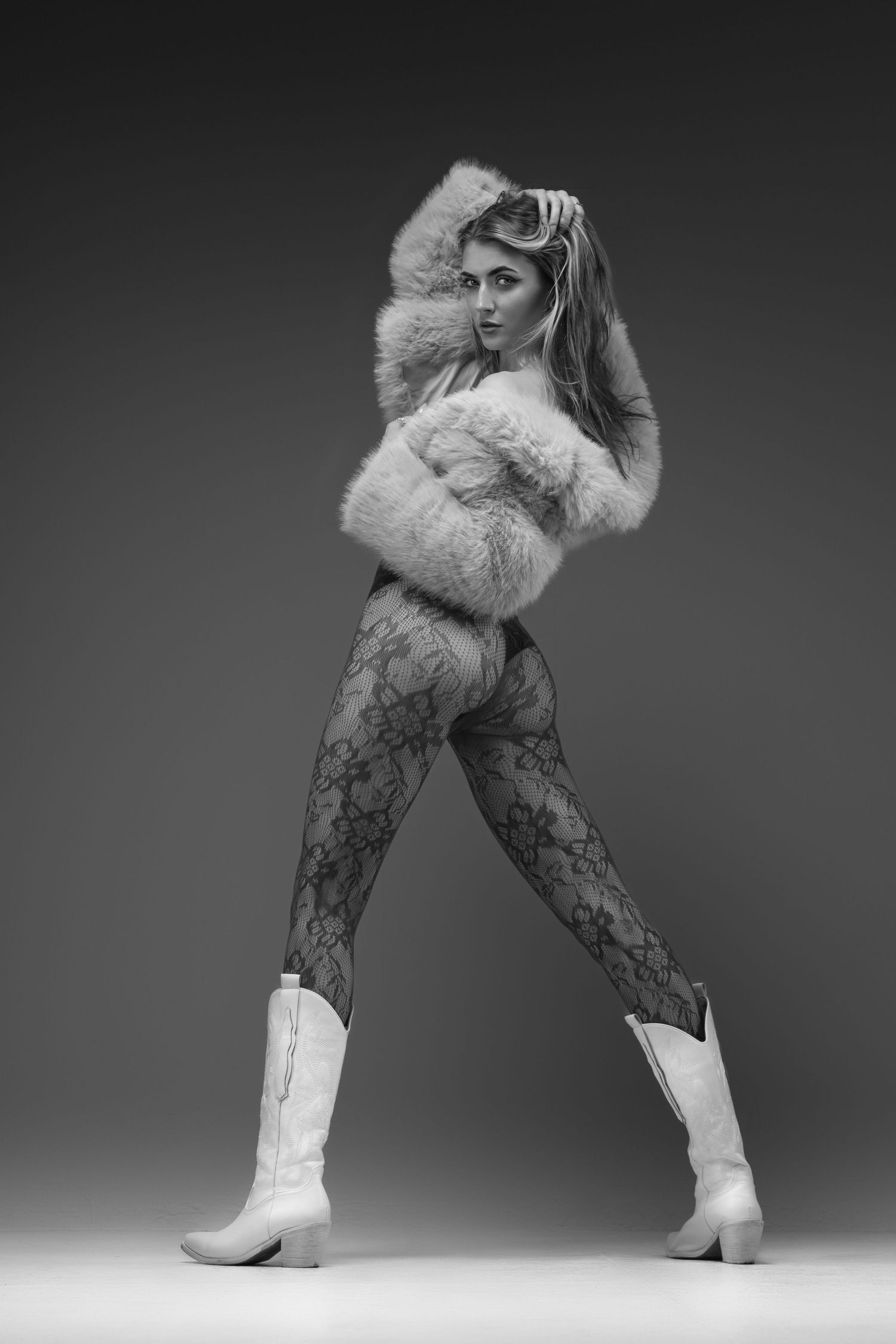 Black and white photo of a woman in a fur jacket, lace tights, and white cowboy boots posing in a studio.