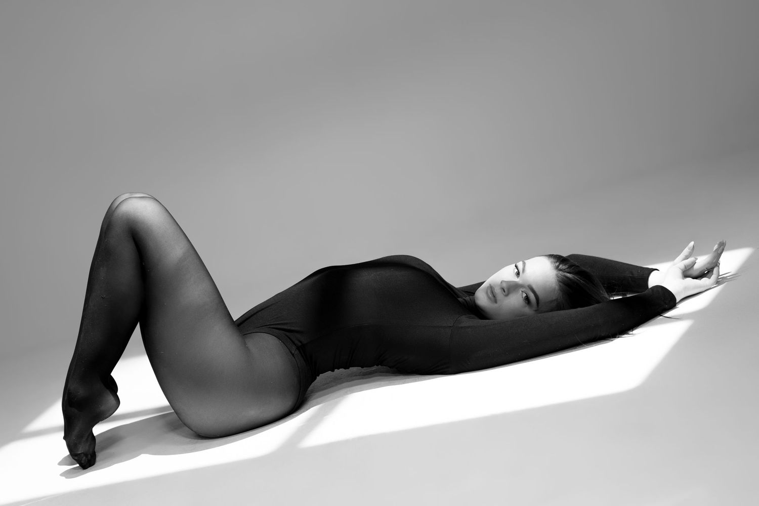 Black and white photo of a woman in a black bodysuit posing gracefully on the floor in dramatic lighting.