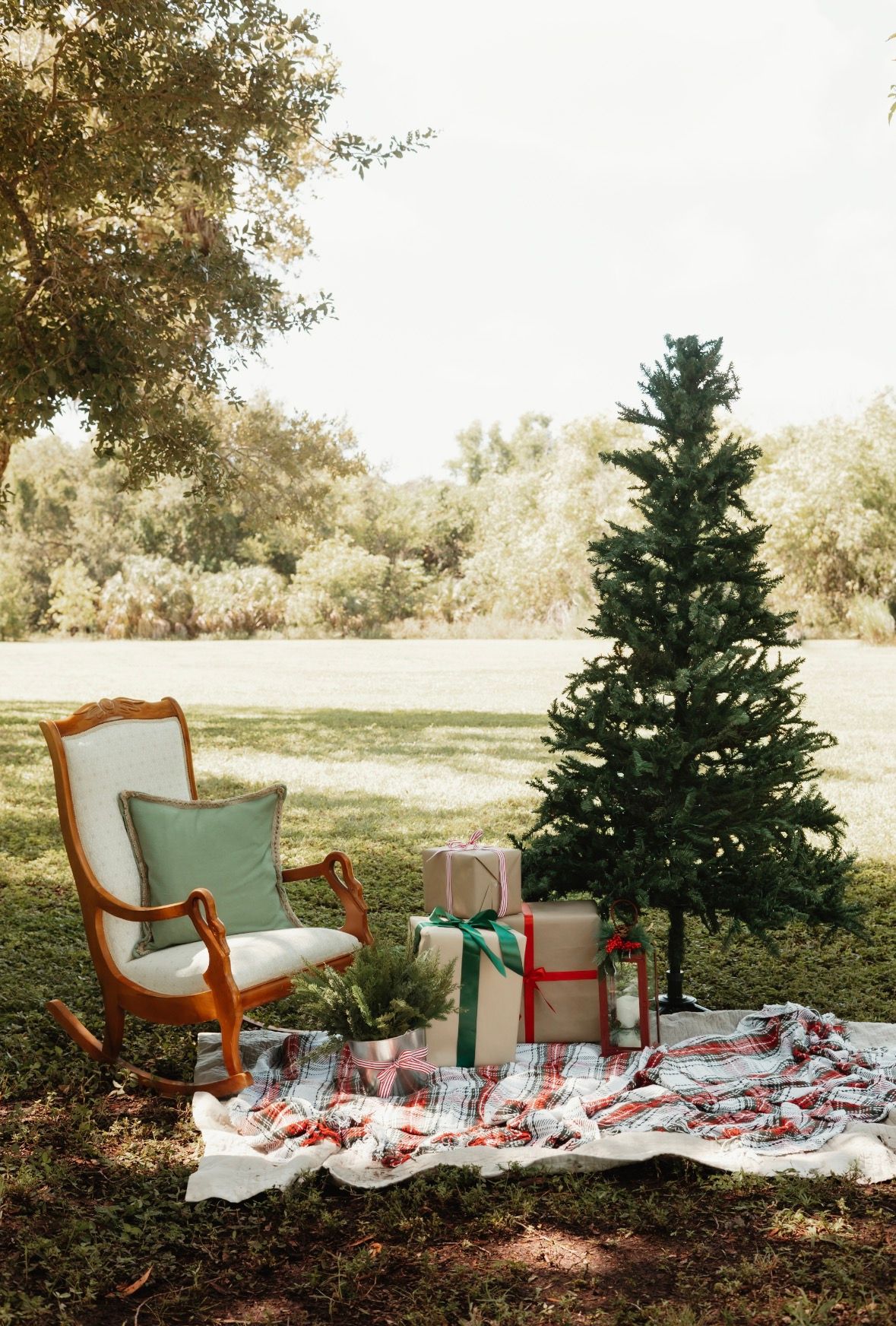 Outdoor Christmas scene with a vintage rocking chair, wrapped gifts, and a decorated tree on a plaid blanket in a green park.
