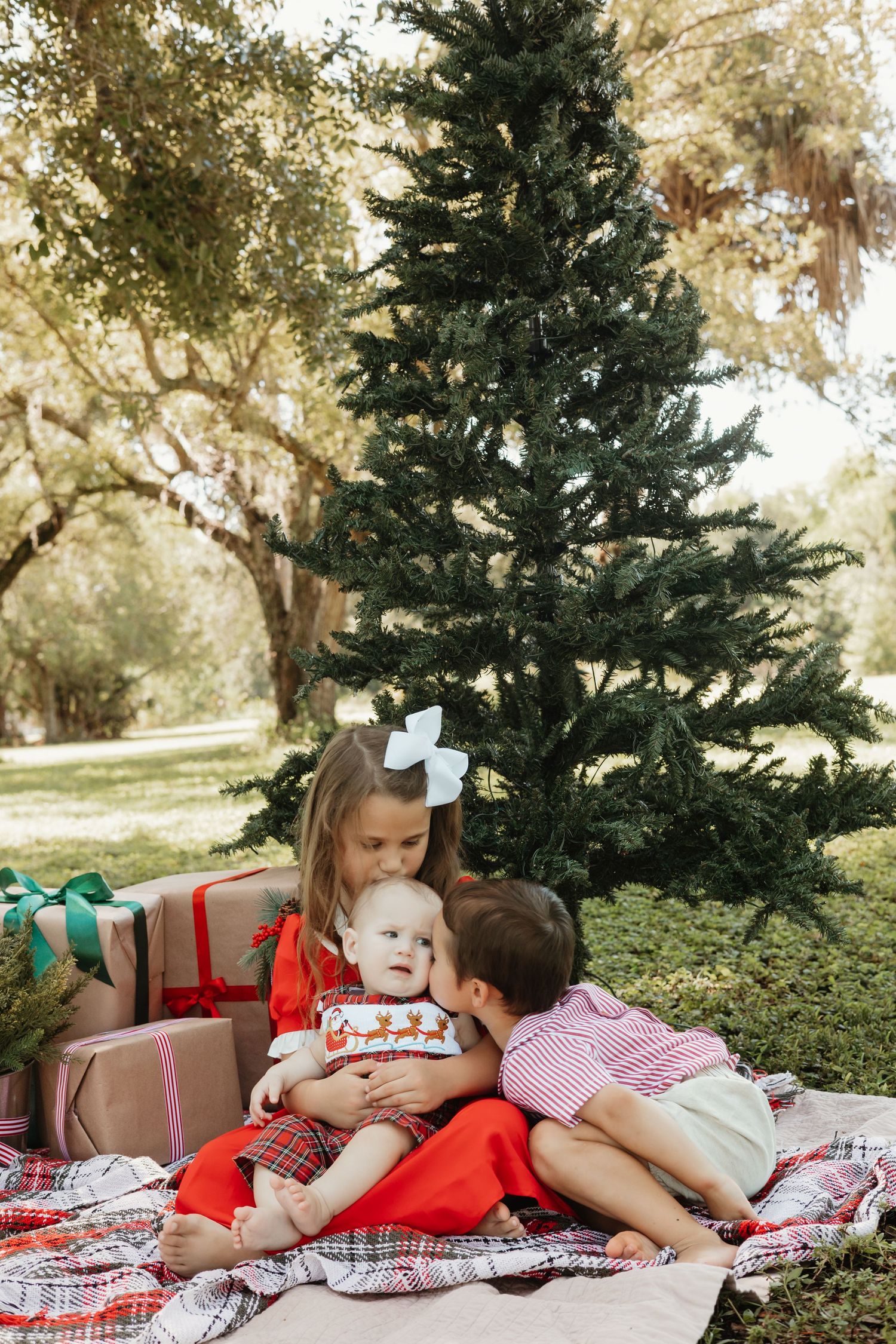 Three children sit on a plaid blanket near a Christmas tree with wrapped gifts outdoors.