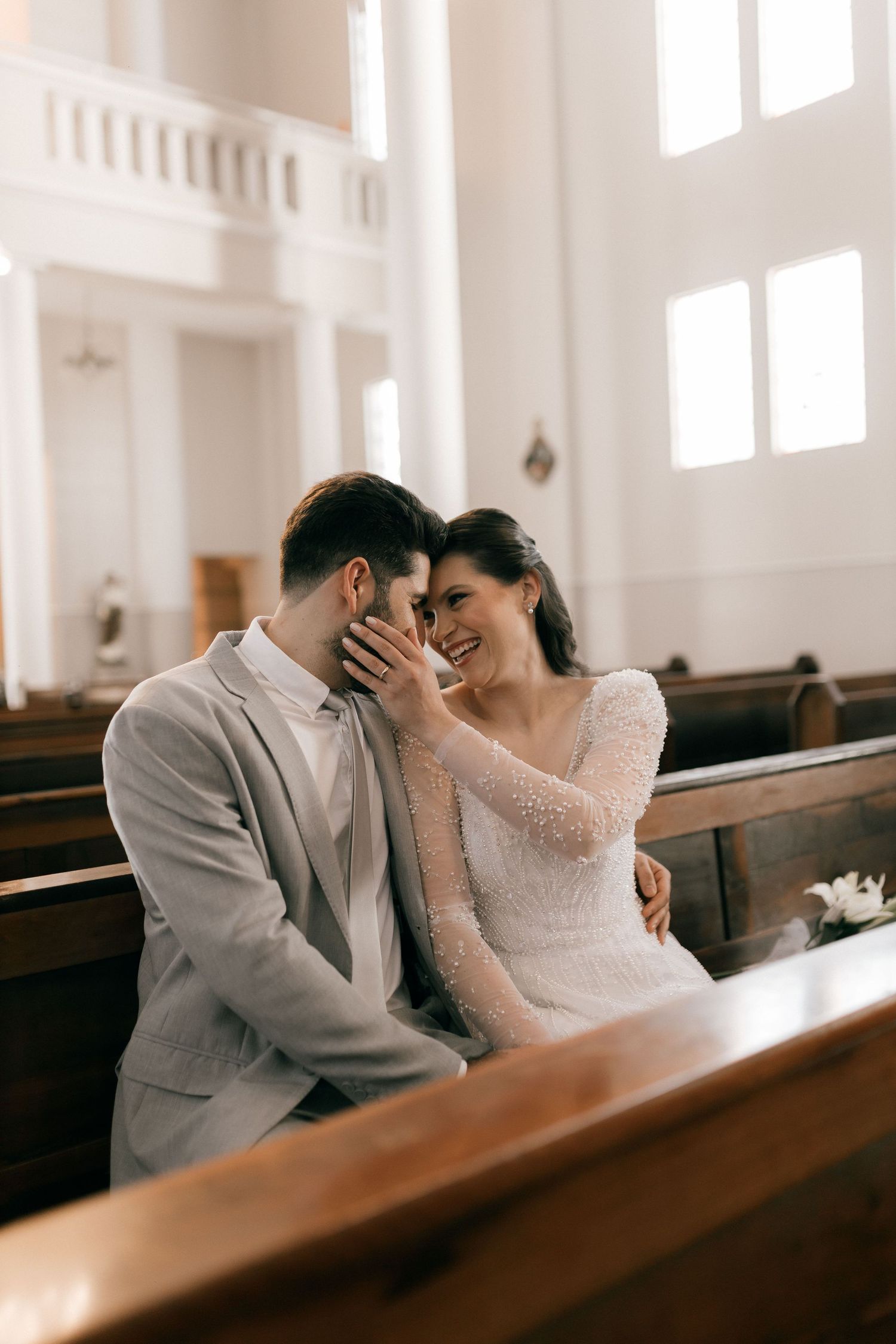 A bride and groom share a tender moment sitting together on a wooden church pew during their wedding ceremony.