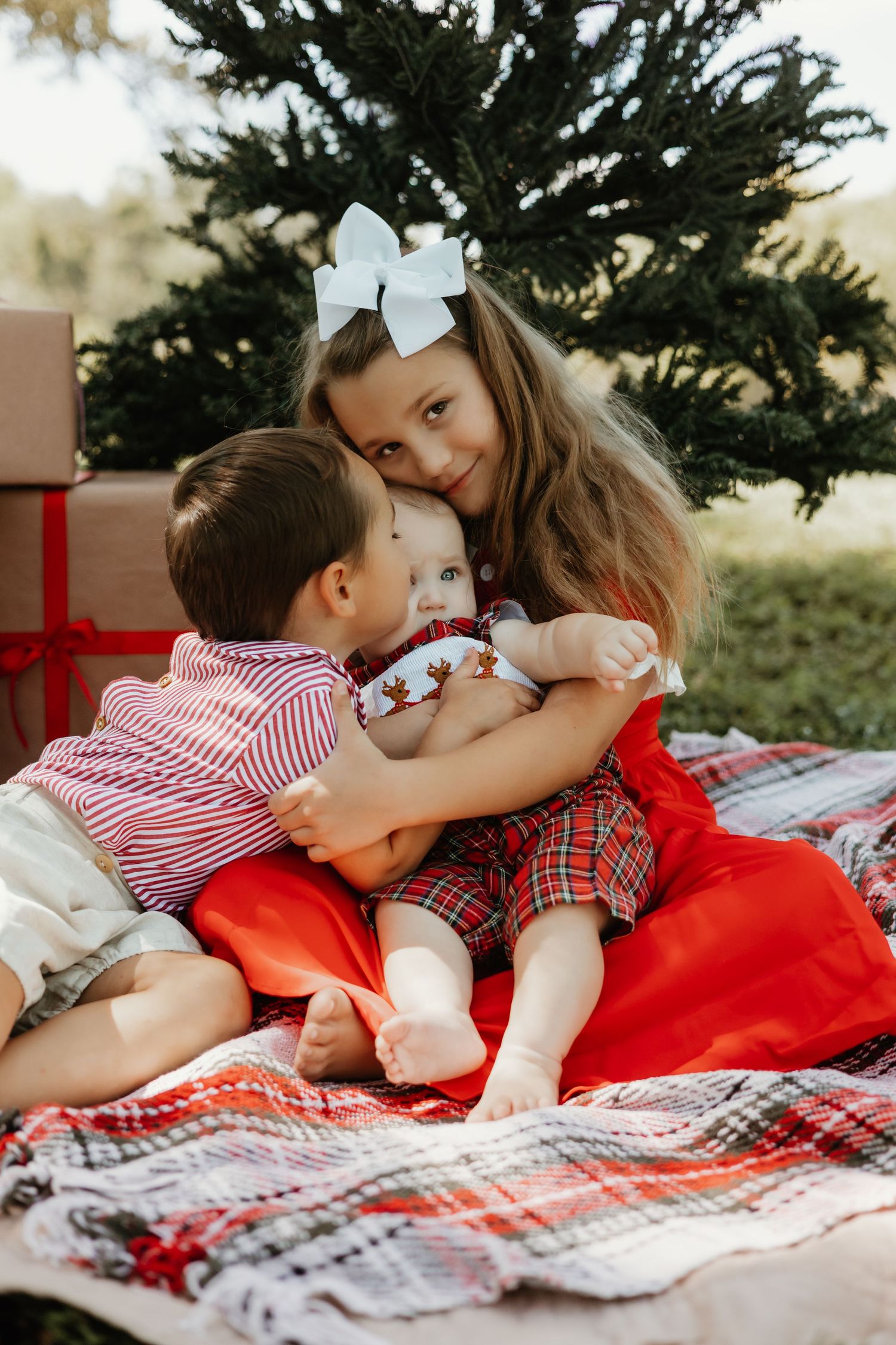 Three children in festive holiday outfits sit together on a plaid blanket near a Christmas tree, sharing a warm embrace.